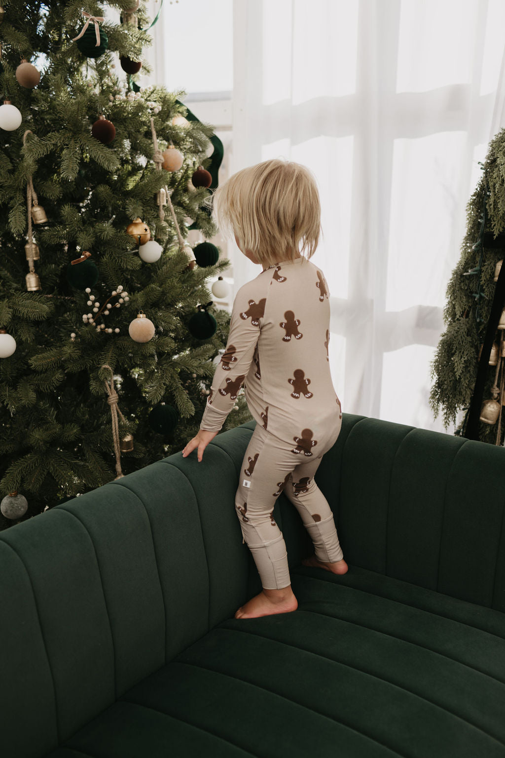 A toddler dressed in forever french baby's Bamboo Zip Pajamas in the Gingerbread pattern stands on a dark green couch, gazing at a decorated Christmas tree. The tree is adorned with various ornaments, and soft light filters through a window with sheer curtains, casting a warm glow over the cozy scene.