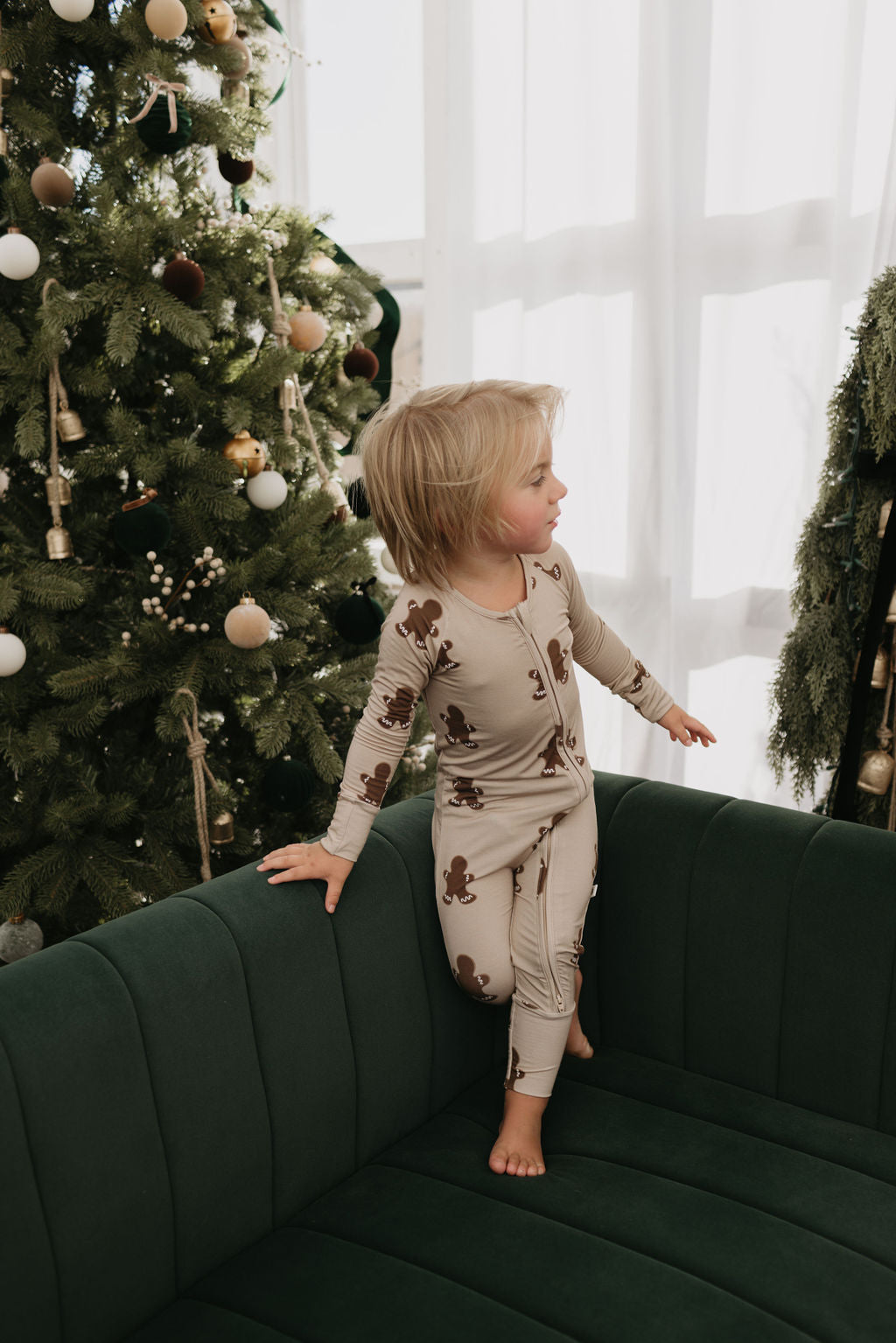 A child wearing the Bamboo Zip Pajamas in Gingerbread by Forever French Baby stands on a green couch, gazing to the side. The room features a Christmas tree decorated with ornaments and a snowy garland, while soft natural light streams through a curtained window.