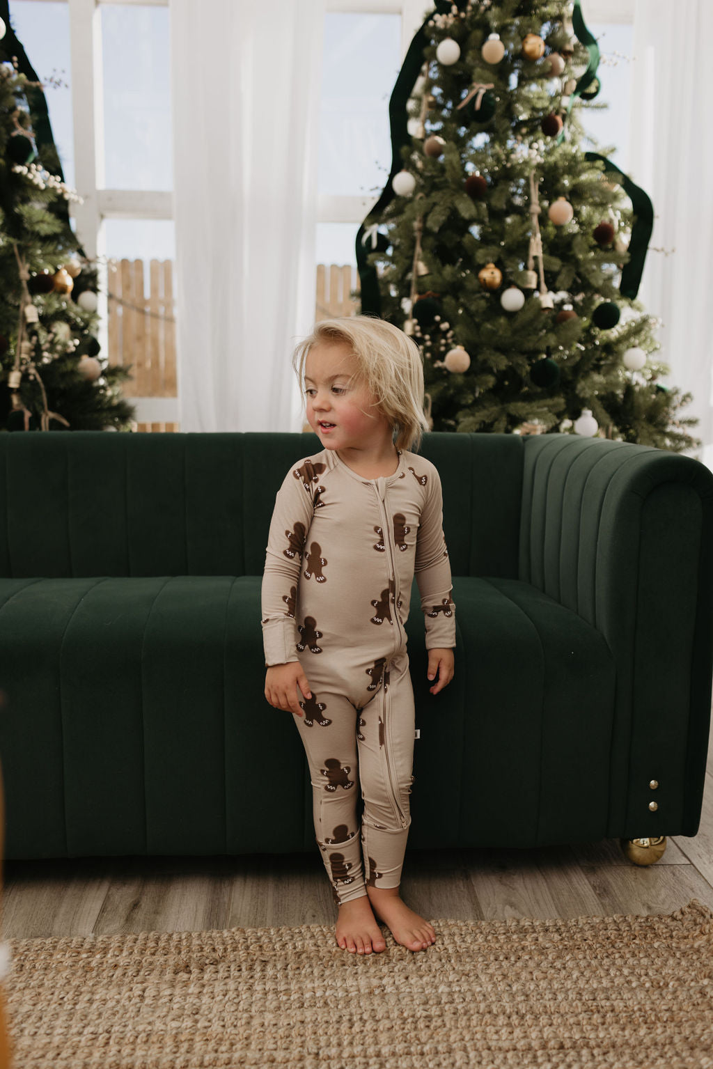 A child in Bamboo Zip Pajamas by forever french baby with a Gingerbread pattern stands beside a green sofa. Christmas trees adorned with ornaments set the background aglow. The setting appears cozy and festive, and the bamboo viscose ensures comfort and hypo-allergenic ease for sensitive skin.