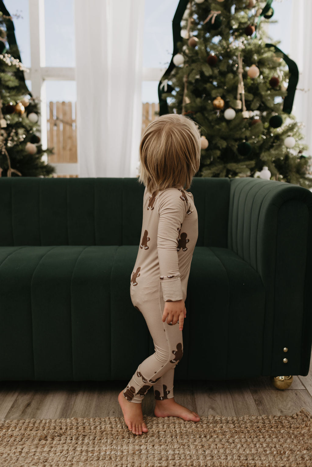 A child dressed in Bamboo Zip Pajamas featuring a gingerbread design from forever french baby stands on a cozy rug, facing sideways, in a living room decorated with Christmas trees and ornaments. The scene is warm and festive, with a dark green sofa nearby.