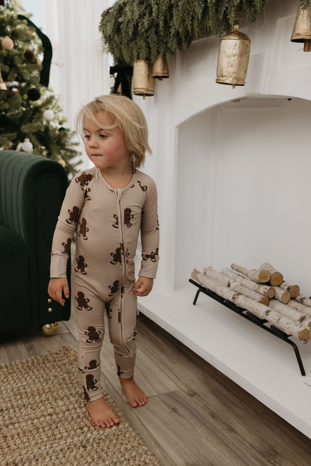 A young child with blonde hair is wearing the cozy Bamboo Zip Pajamas in the Gingerbread pattern from forever french baby, standing near a white fireplace adorned with greenery. Logs are placed in the fireplace, and a Christmas tree glows warmly in the background.