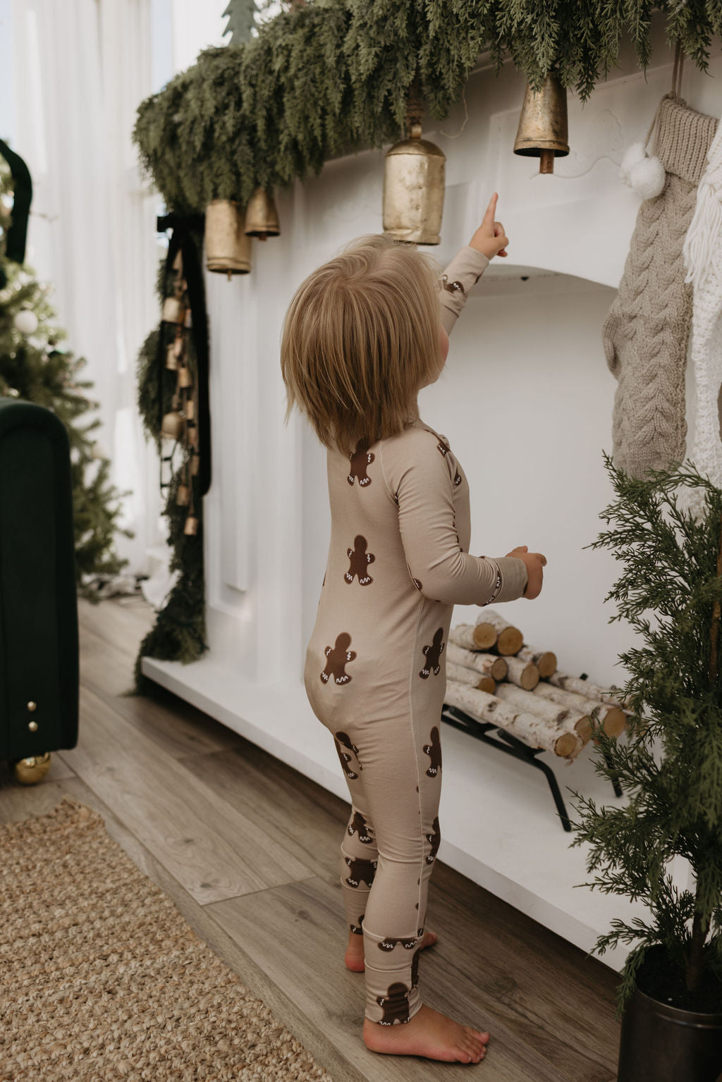 Dressed in forever french baby's Bamboo Zip Pajamas in a charming gingerbread pattern, a child reaches up to touch a bell suspended from the garland adorning the fireplace mantel. The room is elegantly decorated for Christmas with stockings, lush greenery, and a small pile of logs.