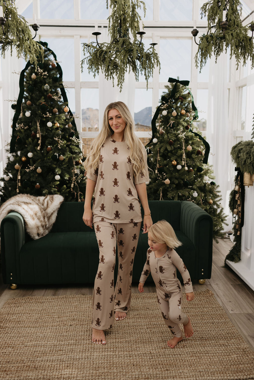 In a cozy room, a woman and a small child are wearing matching forever french baby Bamboo Zip Pajamas in the Gingerbread design. Behind them stand two decorated Christmas trees and a dark green couch, while their hypo-allergenic attire beautifully complements the serene mountain view visible through the glass wall.