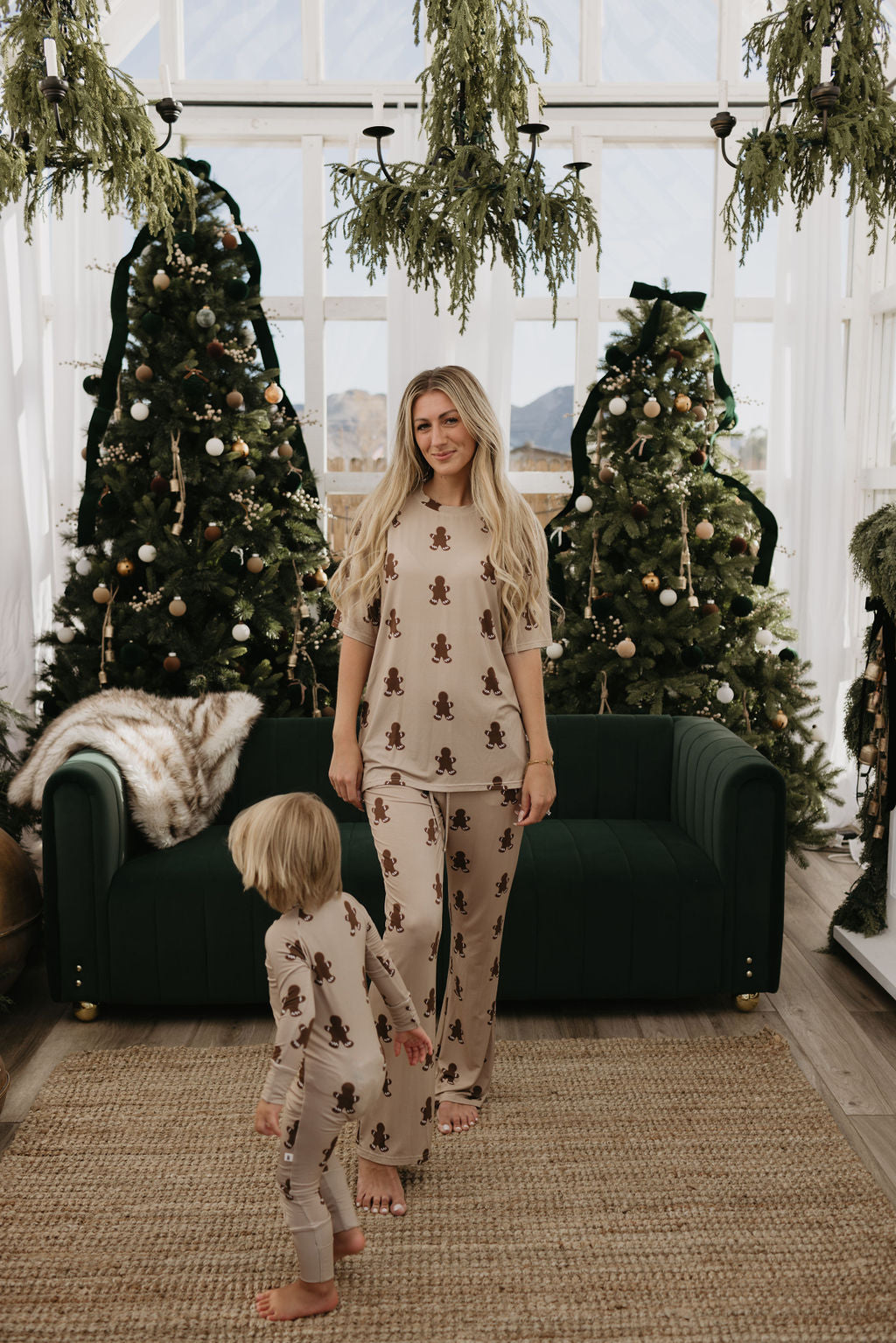 A woman and child in matching Bamboo Zip Pajamas by forever french baby, featuring a gingerbread theme, stand in a festive room adorned with decorated Christmas trees and a dark green sofa. The room's large windows allow natural light to illuminate their cozy, hypo-allergenic clothing amidst the holiday cheer.