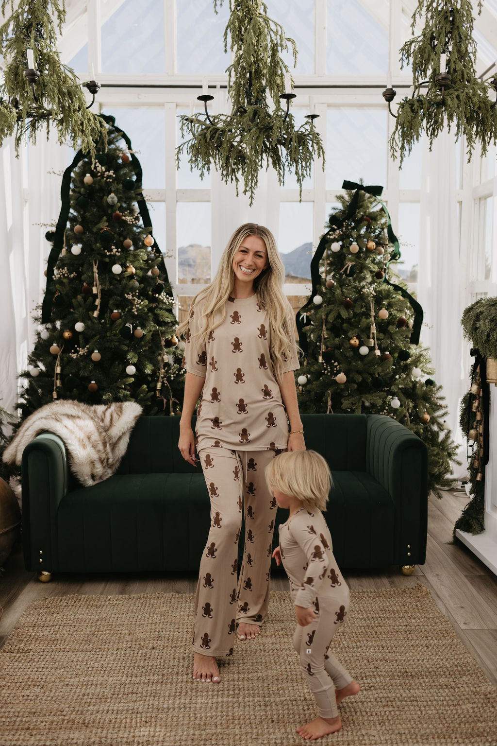 A person and a child stand in front of a dark green couch, dressed in matching oversized Short Sleeve Women's Bamboo Pajamas in a gingerbread theme from forever french baby. Three decorated Christmas trees are visible in the background, set inside a sunlit room with large windows and hanging greenery.