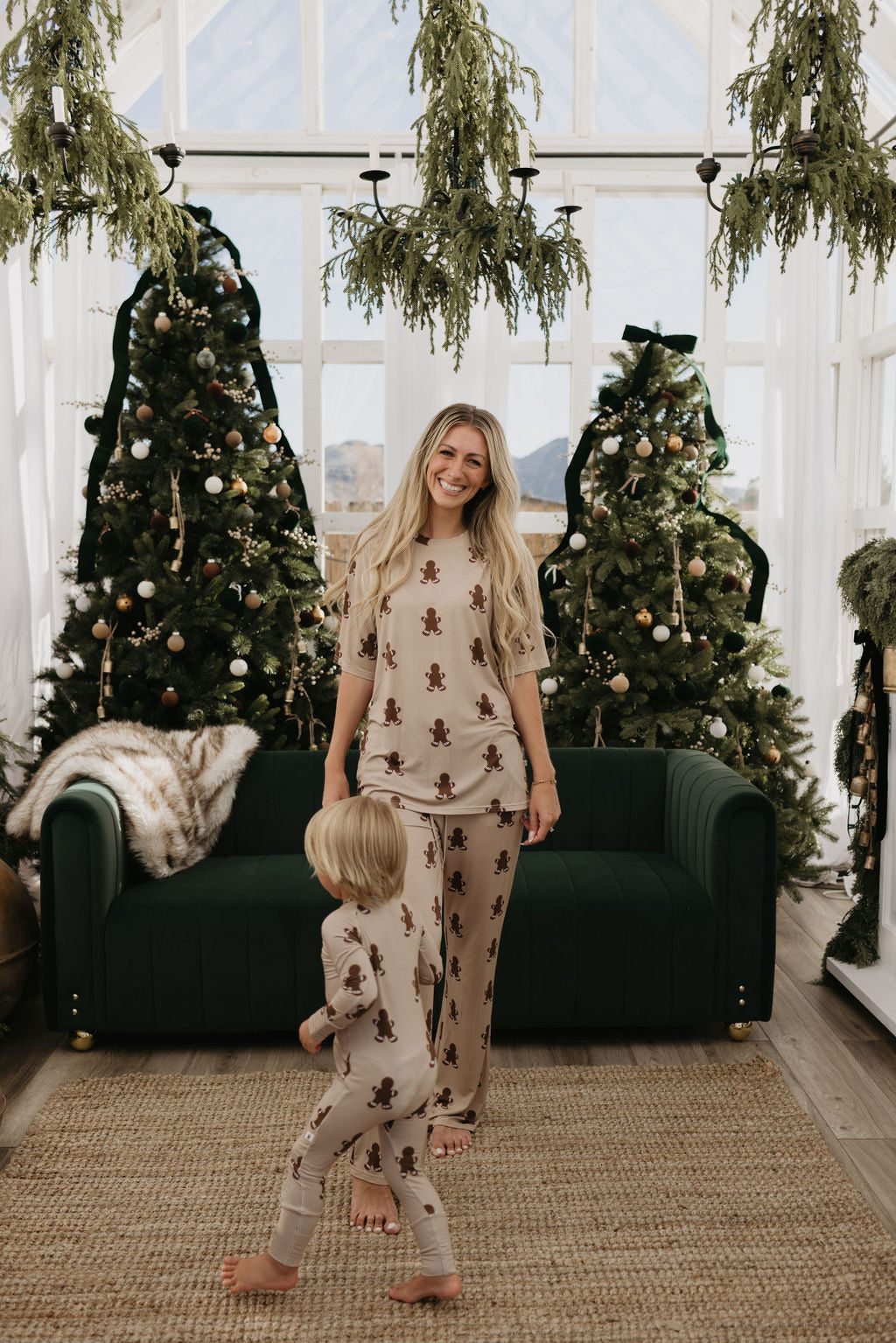 A woman and child, both dressed in Forever French Baby's Bamboo Zip Pajamas featuring gingerbread designs, stand in front of a green couch. Three decorated Christmas trees are visible behind them, and the room's large windows and hanging greenery add to the festive atmosphere.