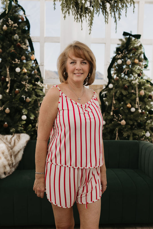 A woman is indoors between two decorated Christmas trees, dressed in a sleeveless, striped red and white top from the "Cami Women's Bamboo Set | the Claus" by forever french baby, along with matching shorts. Nearby is a green couch positioned in front of large windows.