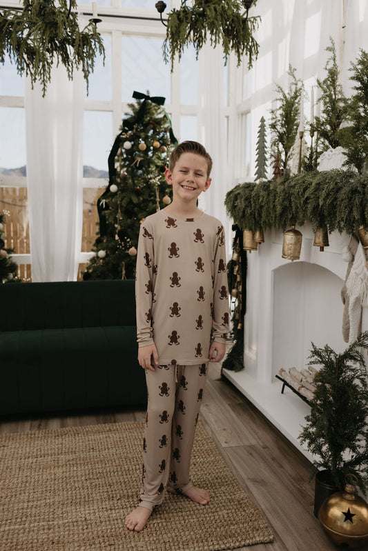 A smiling child stands barefoot indoors wearing forever french baby's Pre-Teen Straight Leg Bamboo Pajamas in a charming gingerbread pattern. Behind them, a decorated Christmas tree and a fireplace adorned with greenery and gold accents create a cozy holiday atmosphere perfect for the hypo-allergenic season.