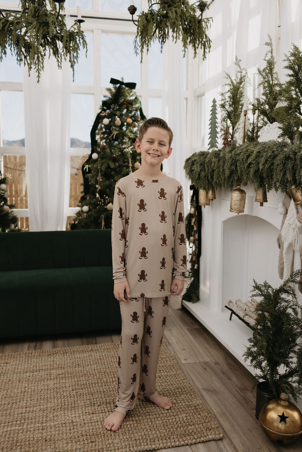 A cheerful child wearing forever french baby's Pre-Teen Straight Leg Bamboo Pajamas in a gingerbread theme stands in a festive room adorned with Christmas trees, greenery, gold ornaments, and stockings hung by a white fireplace.