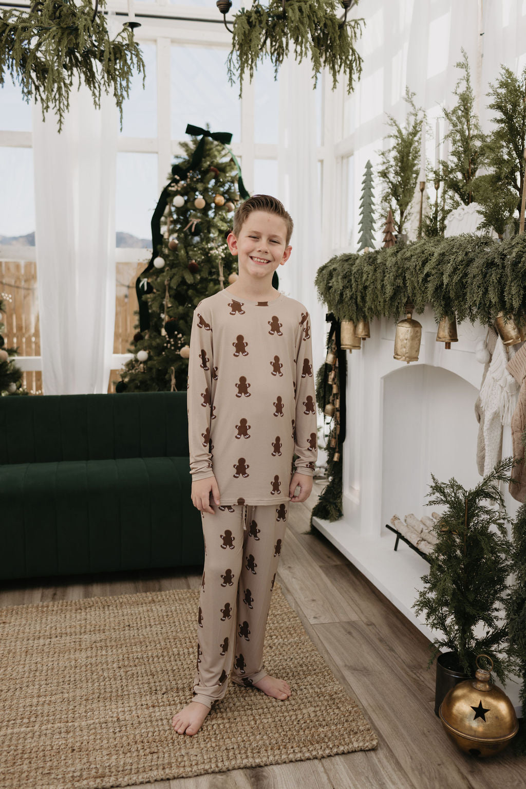 A child is dressed in the Pre-Teen Straight Leg Bamboo Pajamas in Gingerbread by forever french baby, smiling cheerfully in a festive living room decked out with lush greenery and a Christmas tree. The room features a dark green couch and a fireplace beautifully decorated with ornaments and plants.