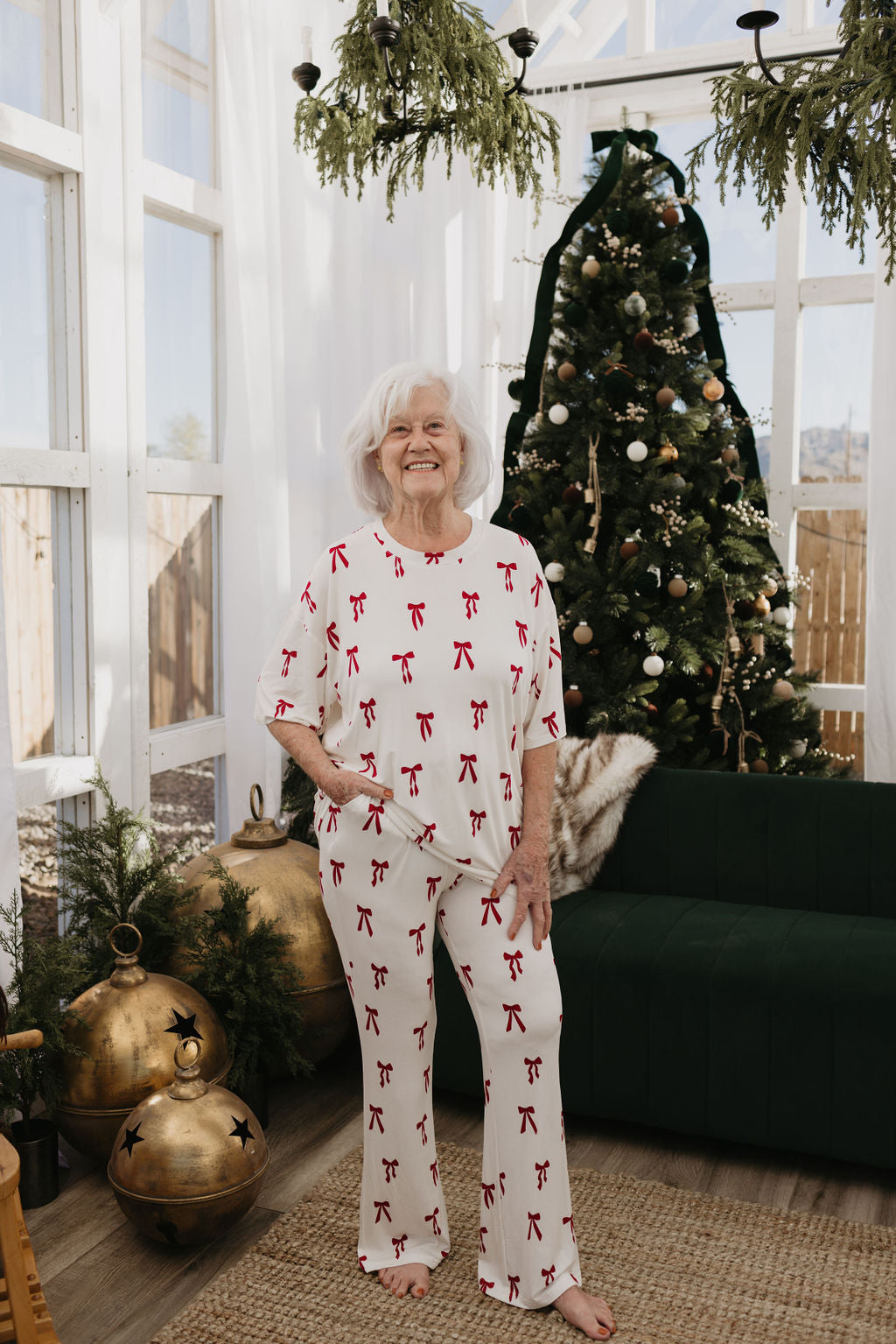 An elderly woman beams in a festive living room, wearing the oversized Short Sleeve Women's Bamboo Pajamas in Ribbons & Bows by forever french baby. Standing beside a decorated Christmas tree and large ornaments, she enjoys the natural light streaming through the glass windows.