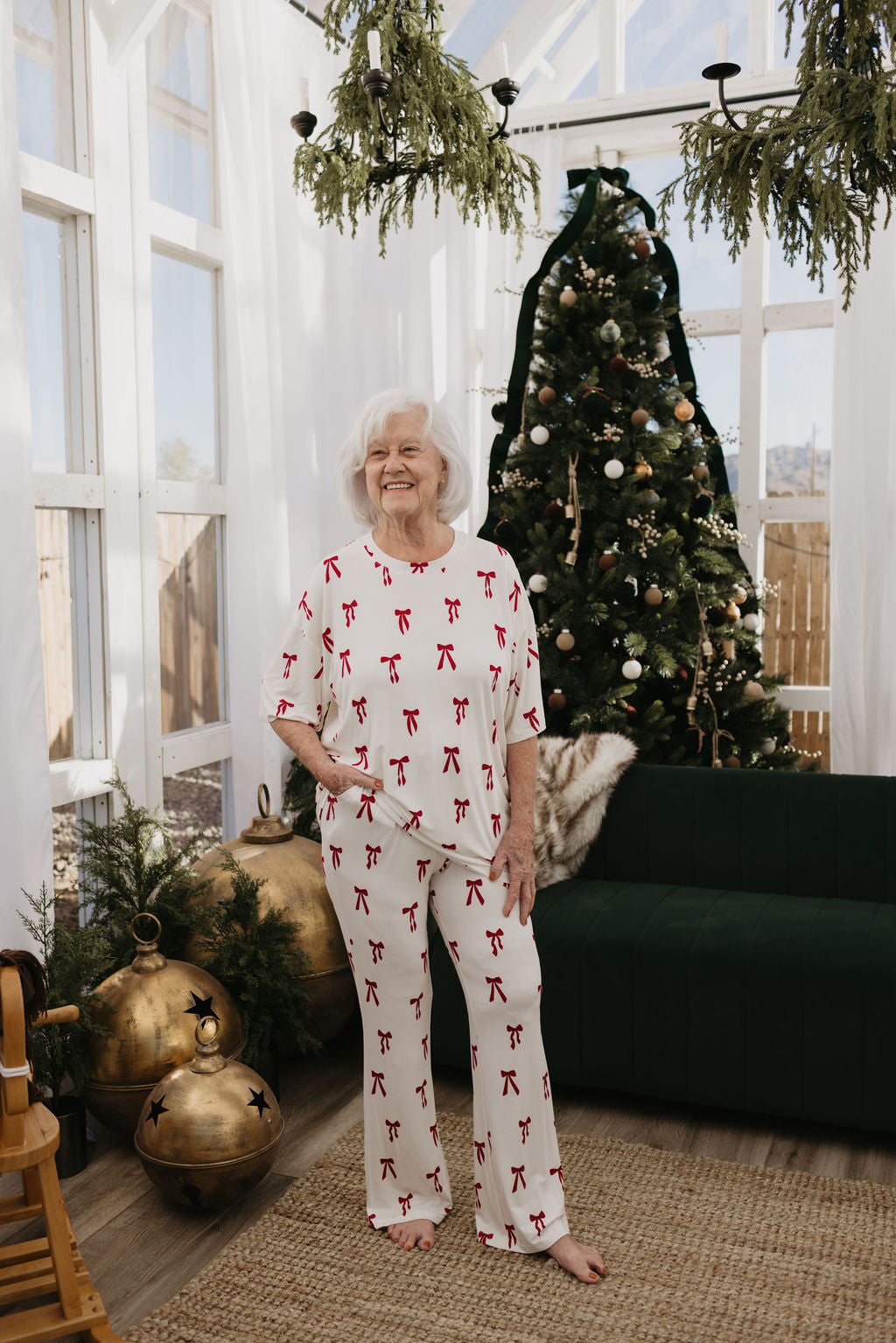 A senior woman with white hair stands smiling in a sunlit room decorated for the holidays. She is wearing an oversized pajama set from forever french baby, featuring their Short Sleeve Women's Bamboo Pajamas in the Ribbons & Bows pattern. A decorated Christmas tree and a green couch create a festive backdrop.