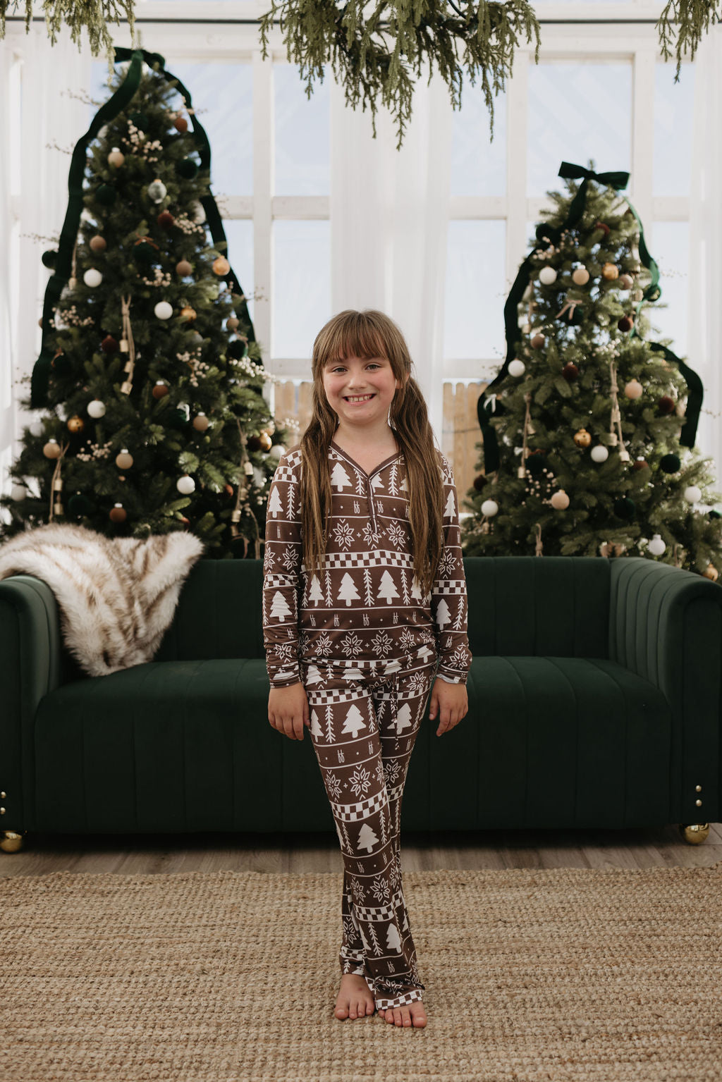 A young girl stands smiling in front of a green couch, wearing the Pre-Teen Flare Bamboo Pajamas in Forever Fair Isle from forever french baby. Crafted from breathable materials, her cozy attire complements the two decorated Christmas trees with ornaments behind her. Natural light filters through the windows, adding to the warmth.