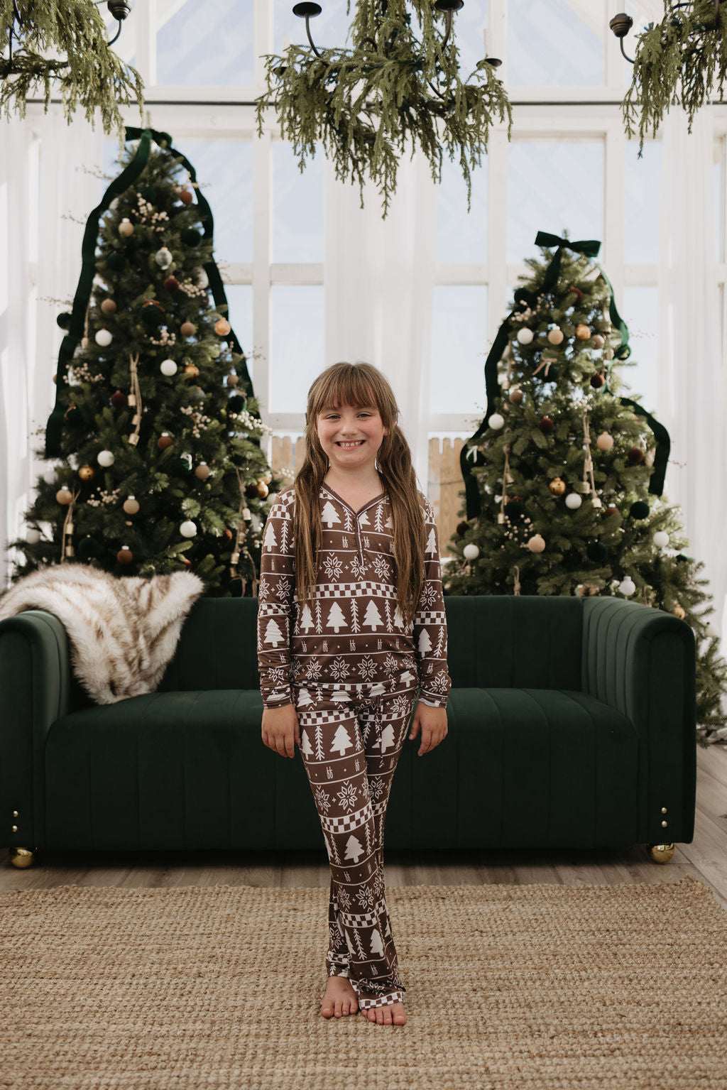 A young girl in "Pre-Teen Flare Bamboo Pajamas | Forever Fair Isle" by forever french baby stands smiling in front of a dark green couch and two decorated Christmas trees, with greenery hanging above. The room is bright with natural light from large windows, accentuating the comfort and style of her cozy hypo-allergenic brown and white holiday pajamas.