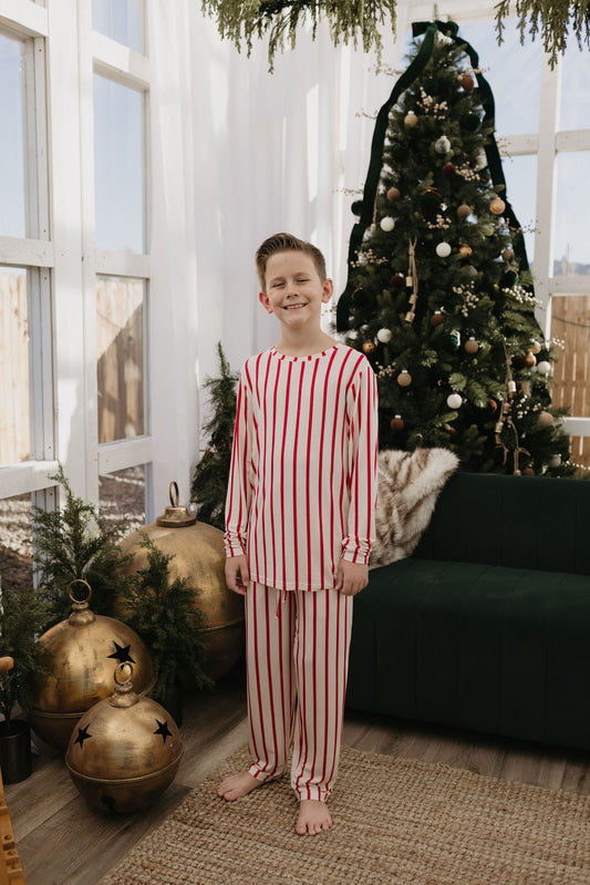 A young boy stands smiling in Pre-Teen Straight Leg Bamboo Pajamas from forever french baby, featuring a red and white striped design. He's in front of a Christmas tree adorned with ornaments, set in a room filled with holiday decorations like gold ornaments and greenery. The pajamas are made from breathable bamboo fabric, providing comfort as he enjoys the festive setting with large windows.