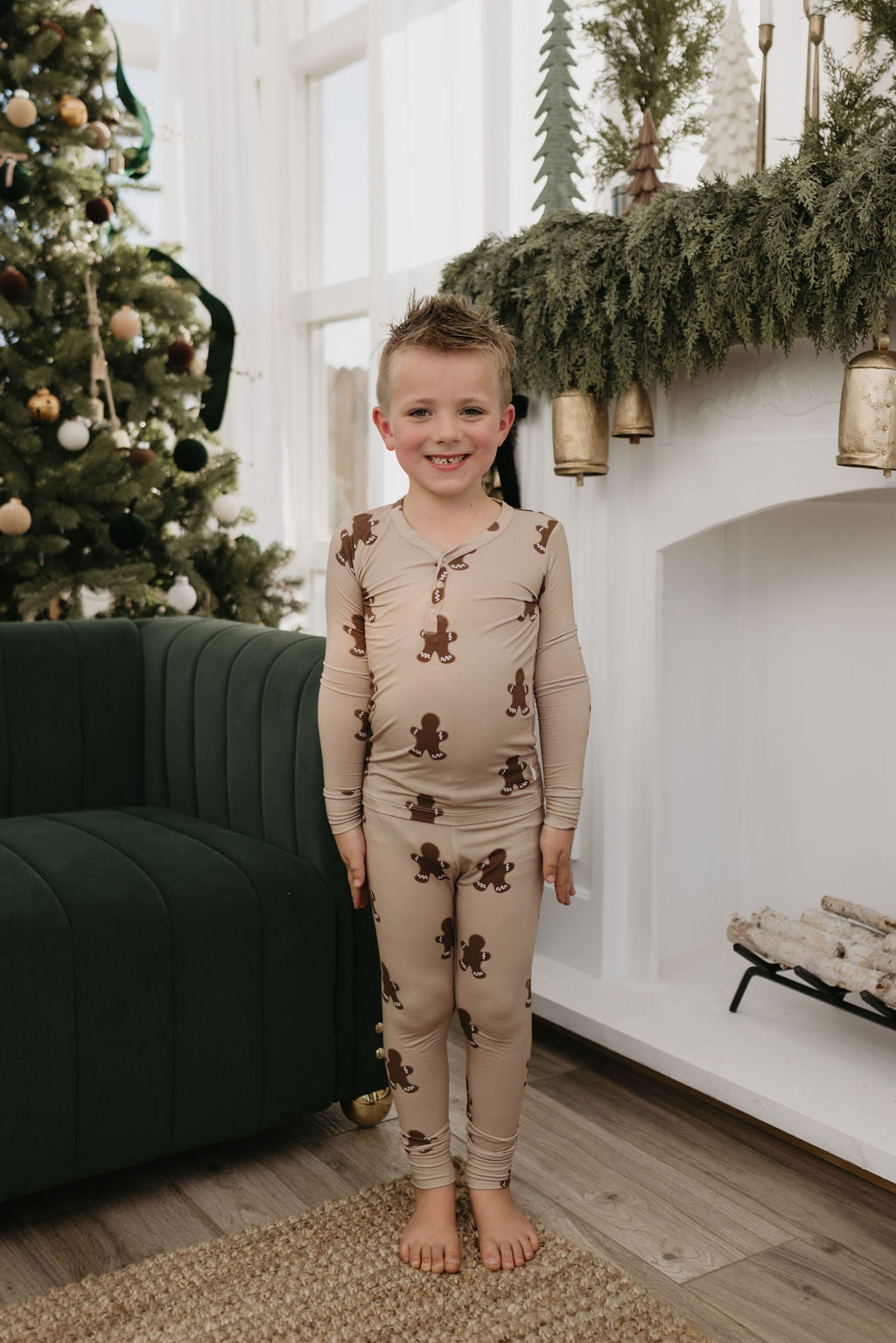 A young boy wearing forever french baby's Bamboo Two Piece Pajamas in a gingerbread theme stands smiling in a living room decorated with a Christmas tree and a fireplace adorned with greenery and bells, creating a warm, festive atmosphere.