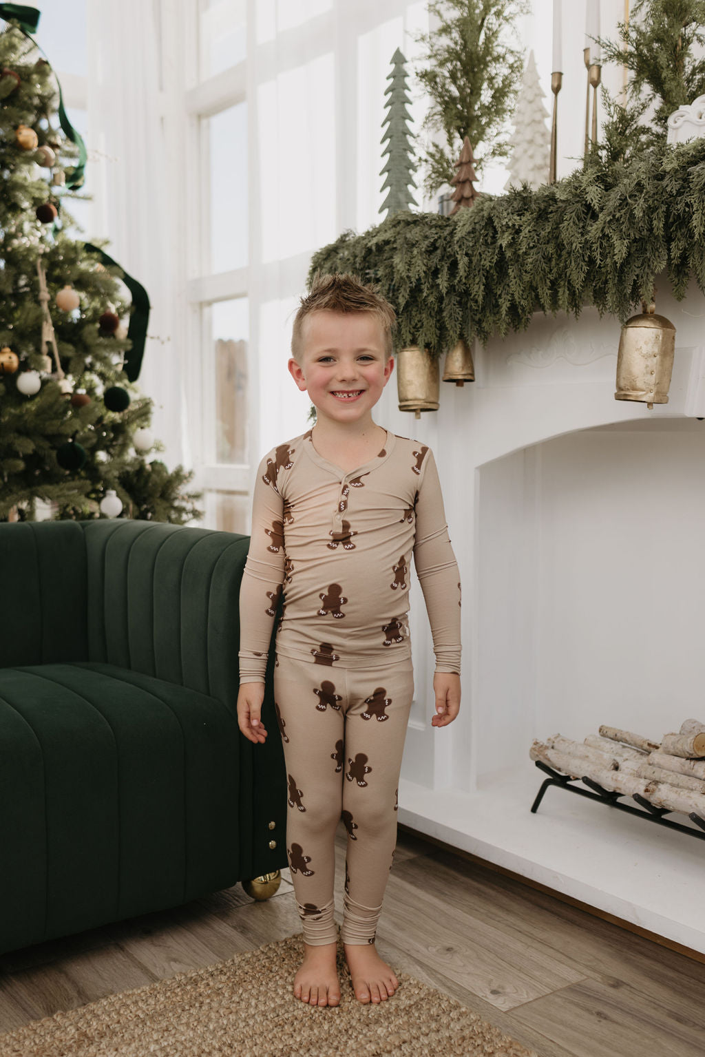 A smiling child stands barefoot in front of a white fireplace decorated with greenery, wearing the forever french baby Bamboo Two Piece Pajamas in a gingerbread pattern. The hypo-allergenic and breathable fabric ensures comfort as a Christmas tree glimmers on the left, with logs filling the hearth.