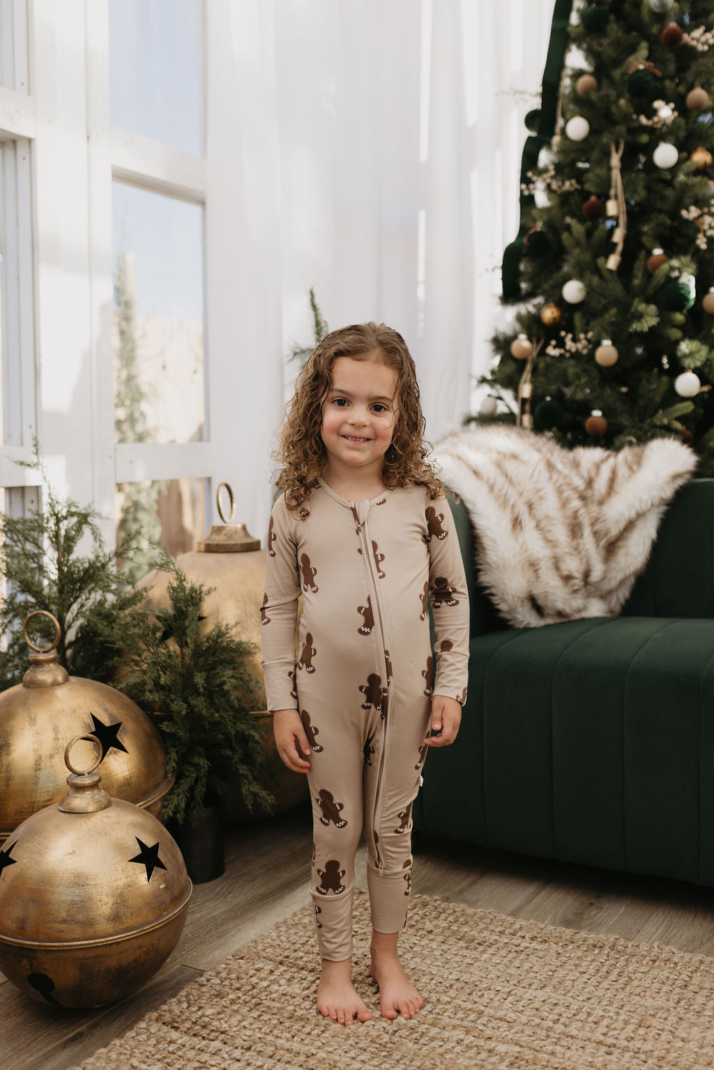 A young child with curly hair stands in a living room wearing the Bamboo Zip Pajamas in a gingerbread pattern from forever french baby. A decorated Christmas tree and large gold ornaments are in the background, creating a festive atmosphere. The hypo-allergenic clothing ensures comfort and joy during holiday celebrations.