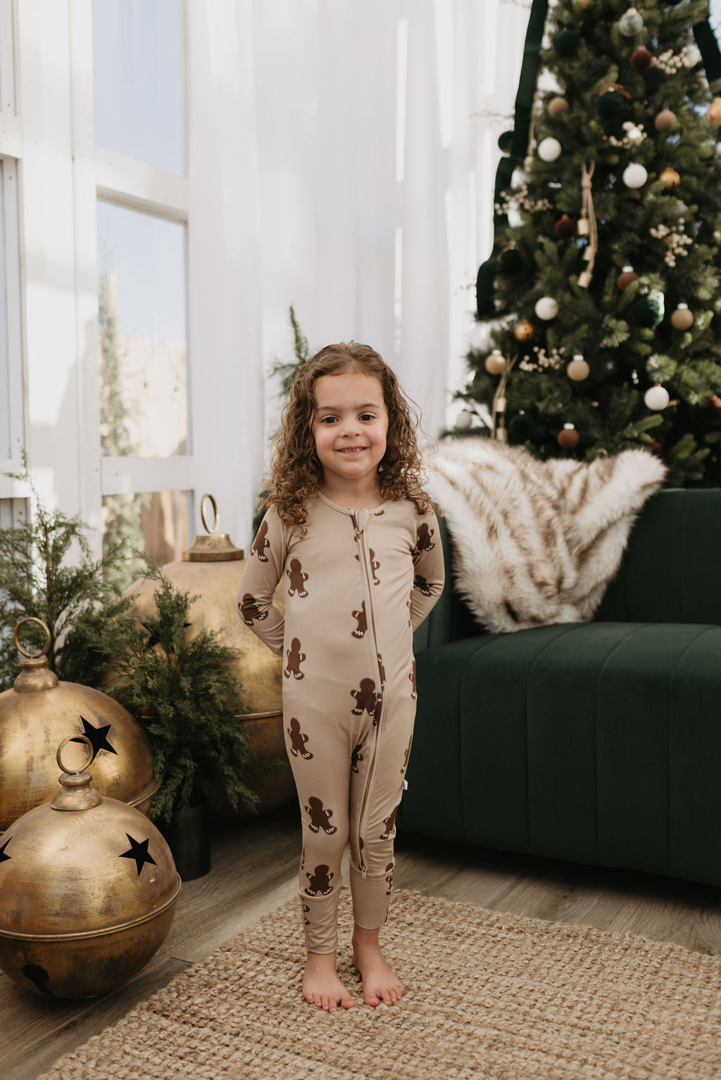 A child with curly hair stands smiling in hypo-allergenic clothing, wearing Bamboo Zip Pajamas in a gingerbread theme by forever french baby. They are beside a green sofa and a decorated Christmas tree with golden ornaments scattered on the floor, creating a cozy room atmosphere.