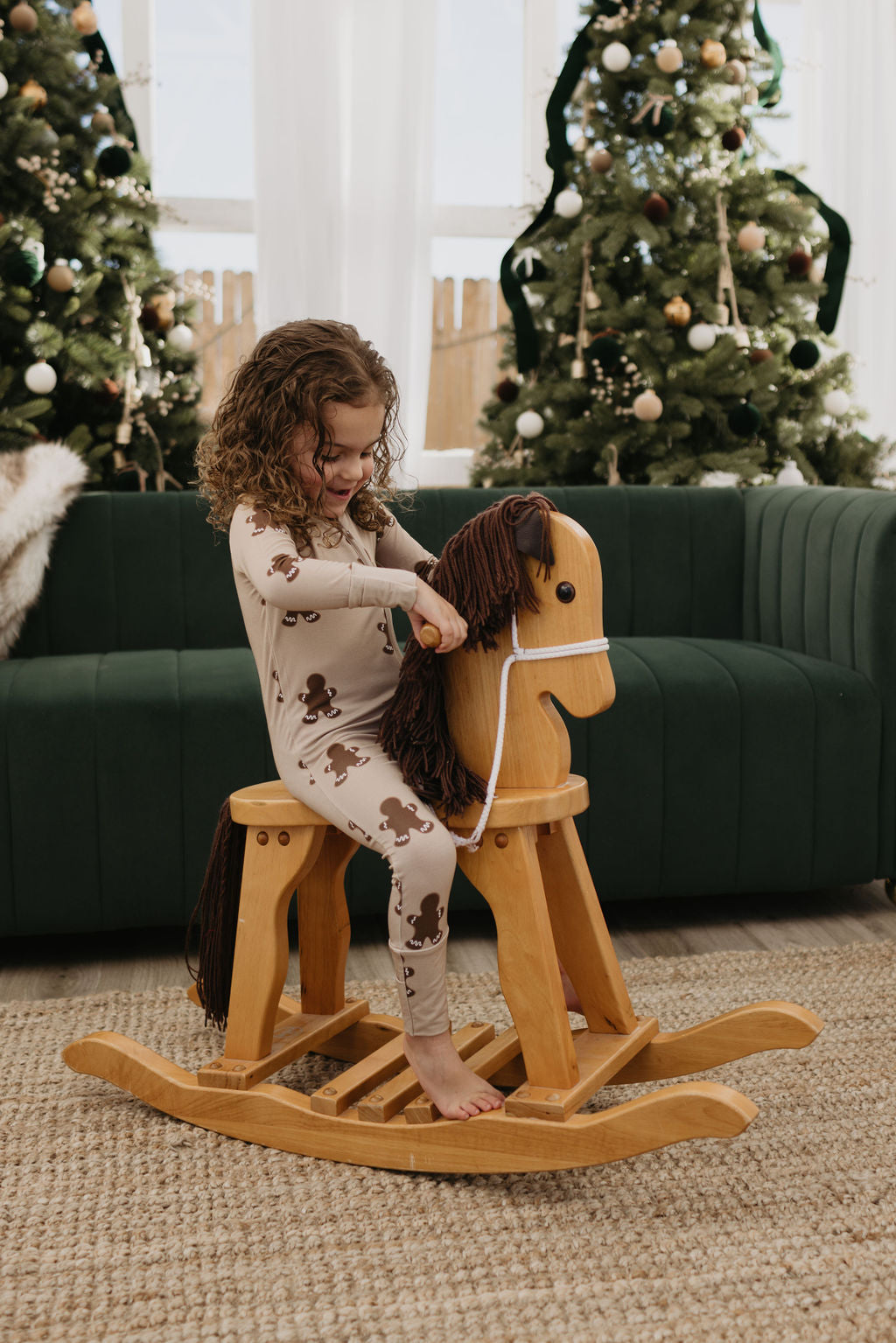 A young child with curly hair is sitting on a wooden rocking horse indoors, wearing cozy bamboo zip pajamas in the Gingerbread design by forever french baby, known for being hypo-allergenic. Two decorated Christmas trees stand in the background next to a green sofa.