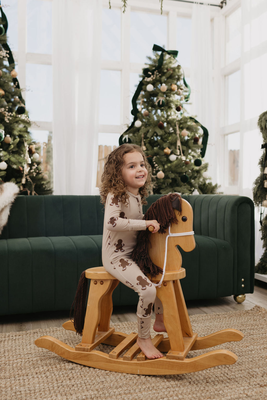 A child with curly hair is grinning on a wooden rocking horse, dressed in Bamboo Zip Pajamas | Gingerbread by forever french baby. The well-lit room, filled with natural light, features two decorated Christmas trees and a green sofa.