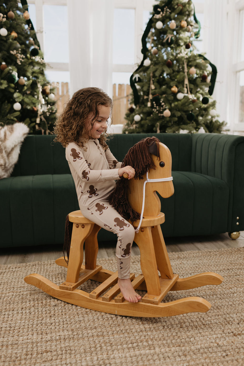 A child with curly hair joyfully rides a wooden rocking horse in the living room. Two decorated Christmas trees are in the background, along with a green couch. The child is wearing Bamboo Zip Pajamas from forever french baby, adorned with a gingerbread print, perfect for cozy and hypo-allergenic comfort.