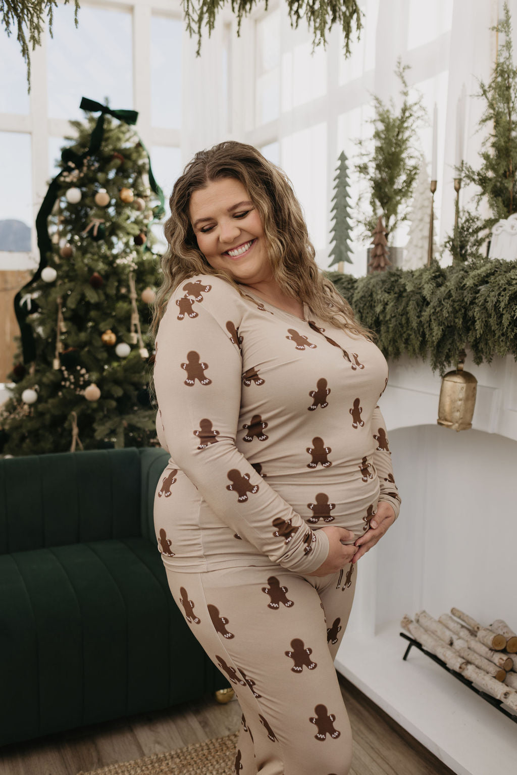 A woman beams with joy, cradling her belly in a warm, festive room. She's dressed in forever french baby's Women's Bamboo Pajamas featuring gingerbread patterns. The room is beautifully adorned with a Christmas tree and garland in the background.