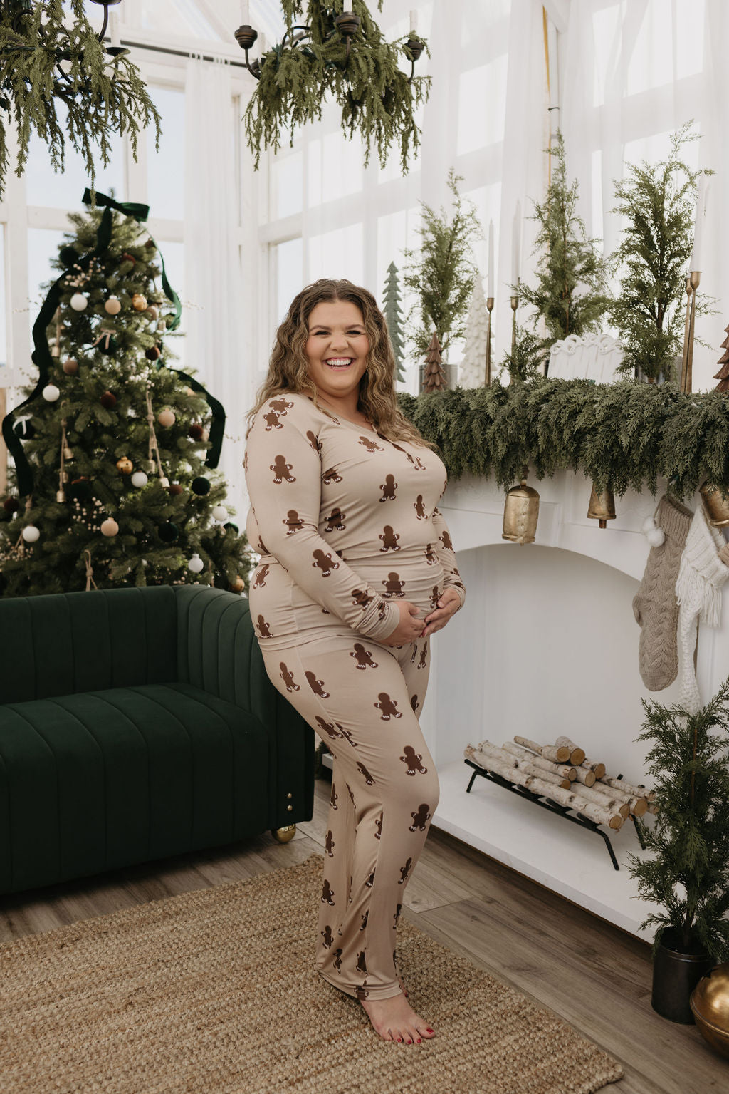 A person dressed in the Women's Bamboo Pajamas | Gingerbread by forever french baby stands in a cozy, festive living room adorned for the holidays with a Christmas tree, garlands, and stockings on the mantle. They are smiling and gently touching their stomach.