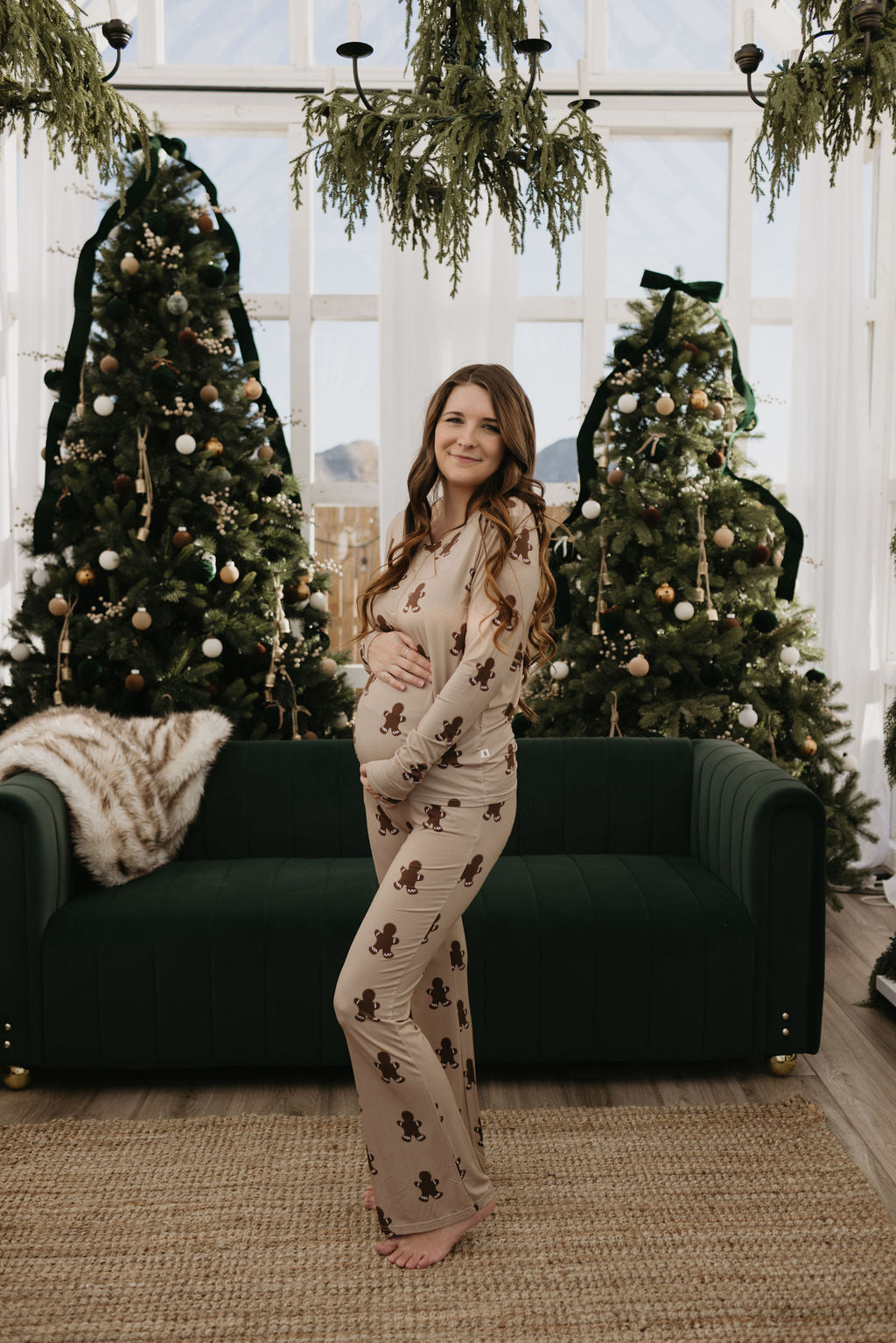 A pregnant woman wearing the Women's Bamboo Pajamas in the Gingerbread pattern by forever french baby stands in front of a dark green sofa. Behind her are two decorated Christmas trees and a window with hanging greenery.