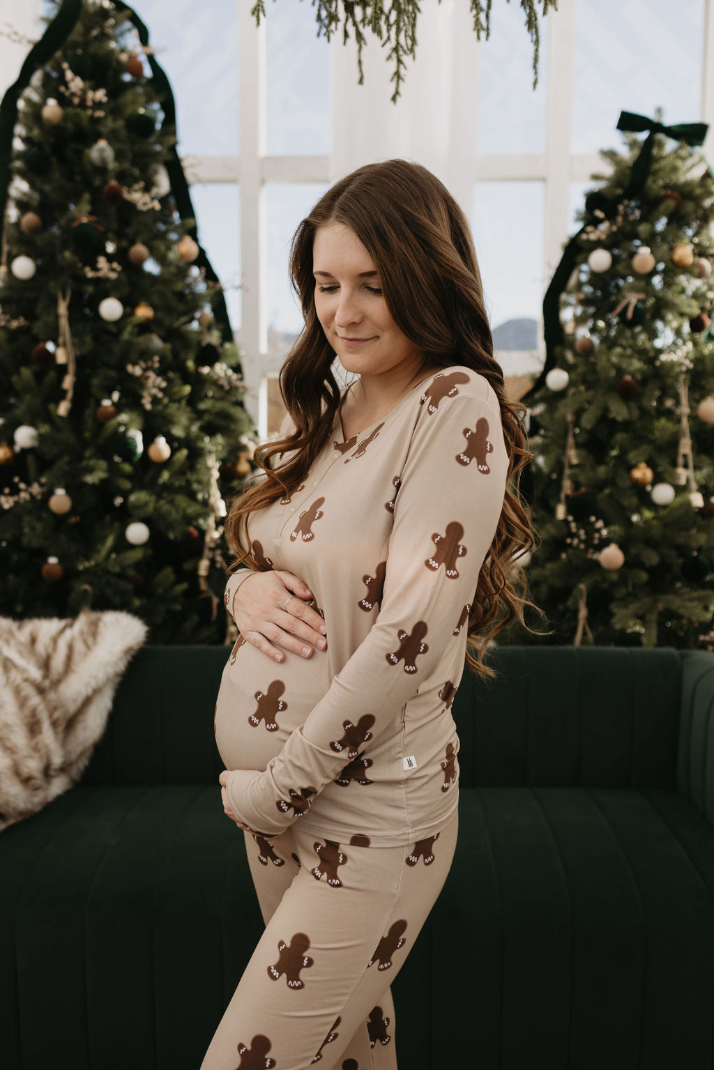 Dressed in forever french baby's Women's Bamboo Pajamas in a gingerbread theme, a woman lovingly cradles her pregnant belly. She stands amidst a festive scene featuring Christmas trees and a dark green couch, embracing the season with cozy comfort.