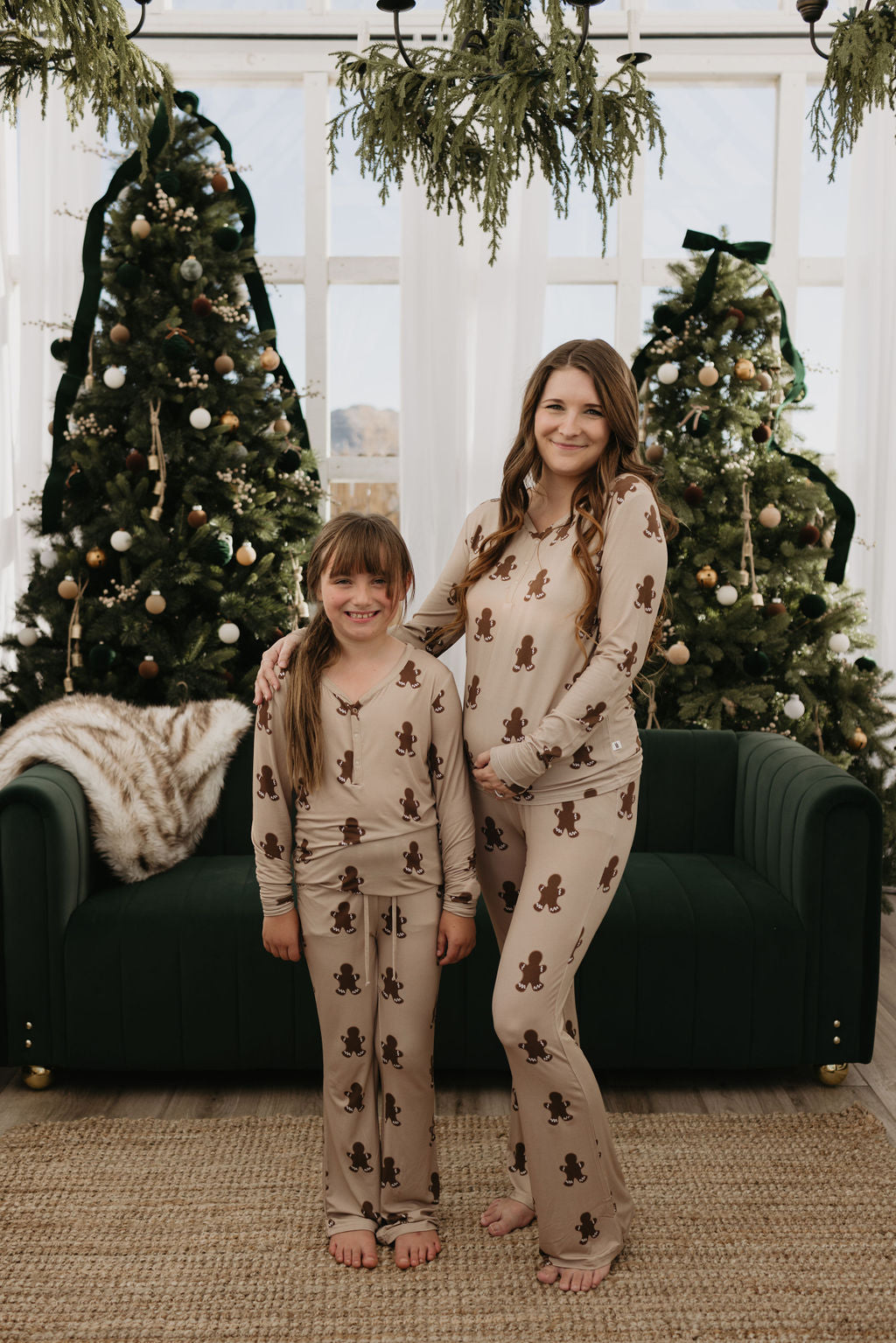 A woman and a girl stand smiling in front of two decorated Christmas trees, both wearing matching pajamas with gingerbread man patterns. Their cozy ensemble includes the Pre-Teen Flare Bamboo Pajamas in Gingerbread by forever french baby, creating a perfect festive look. A green sofa and a fur blanket adorn the background.