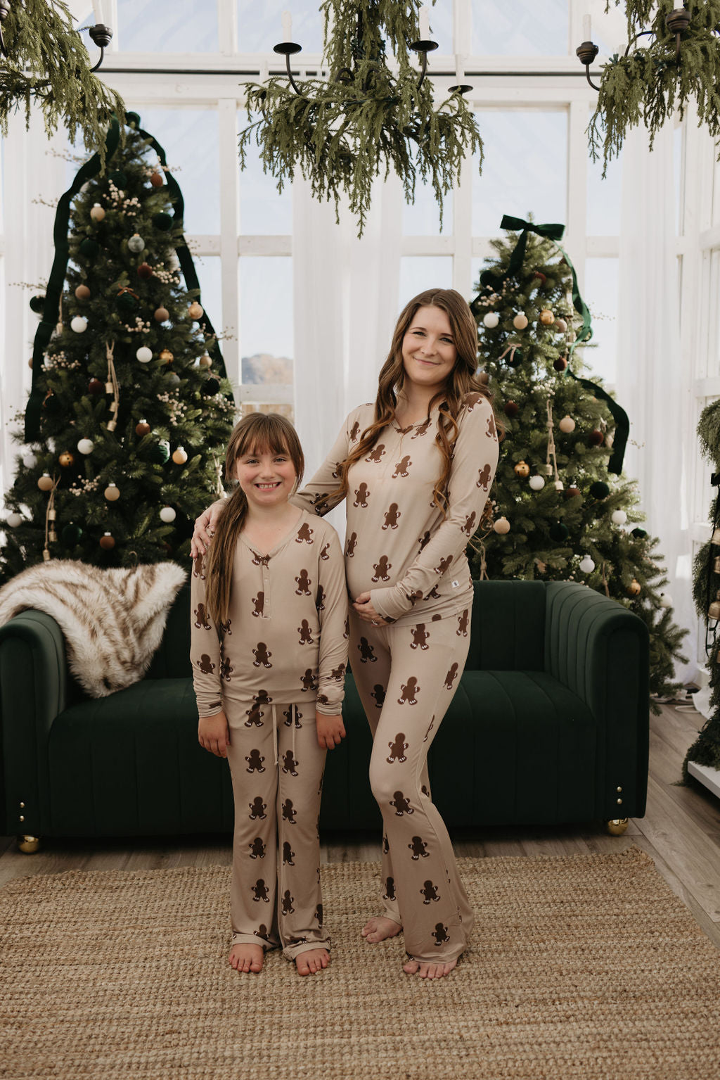 A woman and a pre-teen girl share bright smiles in the cozy living room, wearing matching gingerbread-themed pajamas from forever french baby. The pre-teen sports the Pre-Teen Flare Bamboo Pajamas in Gingerbread. Surrounded by two Christmas trees, a green sofa, and festive greenery, their love for bamboo clothing adds even more warmth to the scene.