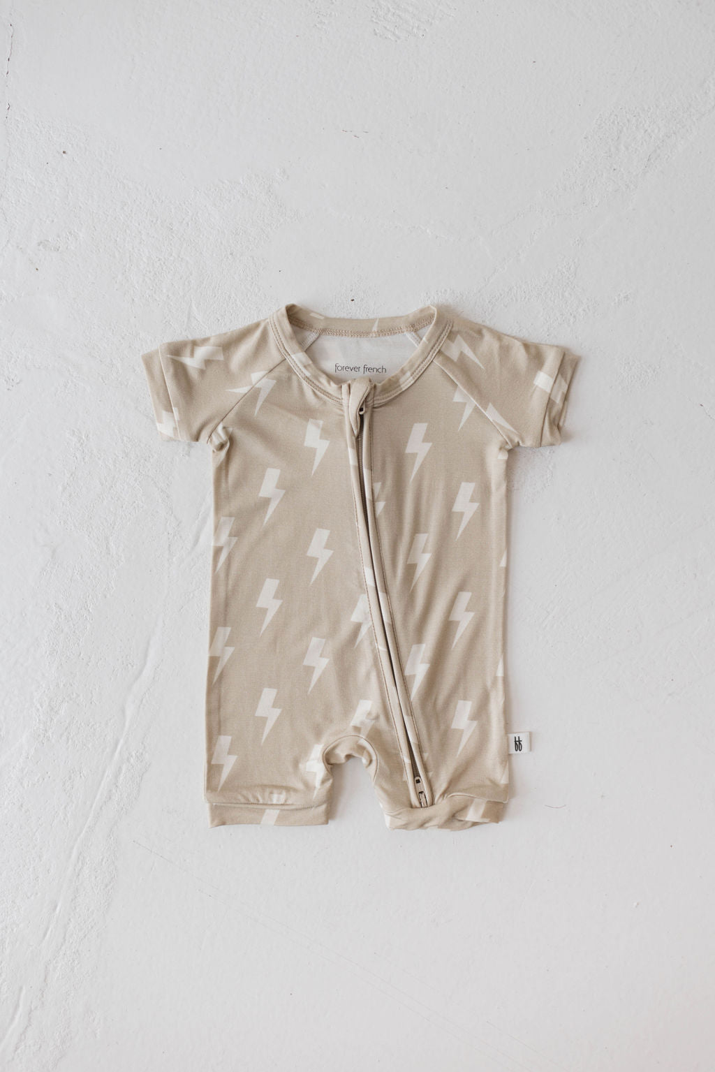 The Shortie Bamboo Zip | Tan Lightning Bolt romper by forever french baby is a small, beige baby outfit featuring short sleeves and legs, adorned with a pattern of white lightning bolts. This hypo-allergenic romper is designed with a diagonal zipper from the neckline down to the left leg for easy dressing. It is displayed flat on a light, textured surface.