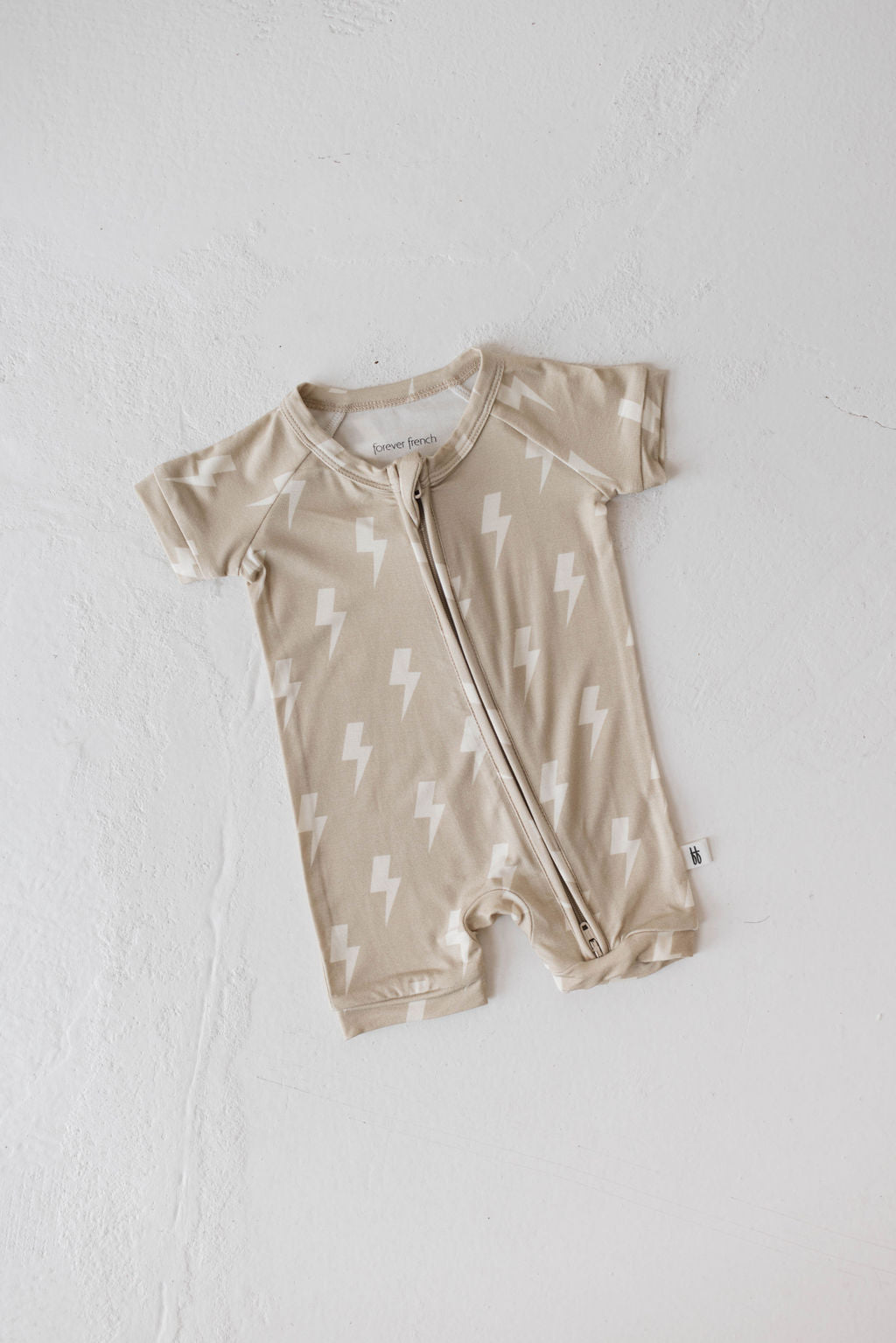 The Shortie Bamboo Zip in Tan Lightning Bolt by forever french baby, a hypo-allergenic beige romper with short sleeves and a front zipper, featuring a pattern of white lightning bolts, is laid flat on a white surface.