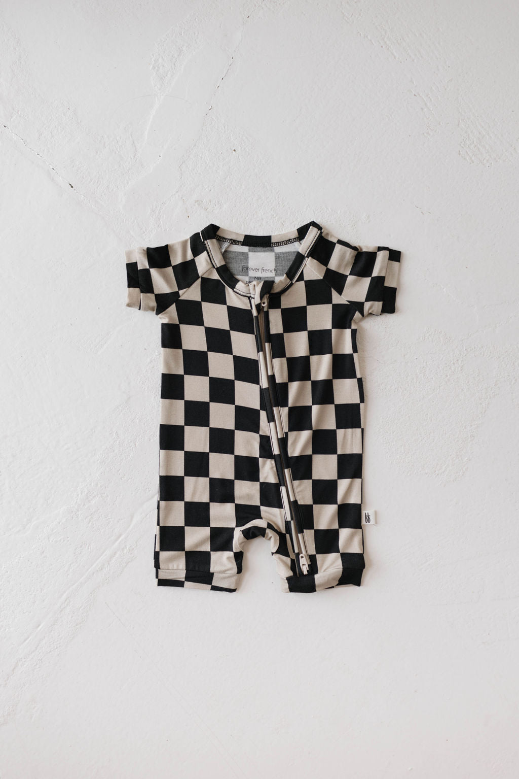 The Shortie Bamboo Zip in Black Checkerboard from forever french baby is laid flat on a light-colored, textured surface. This baby romper, made of hypo-allergenic materials, features short sleeves, short legs, and a front zipper running from the neckline to the crotch for added comfort.