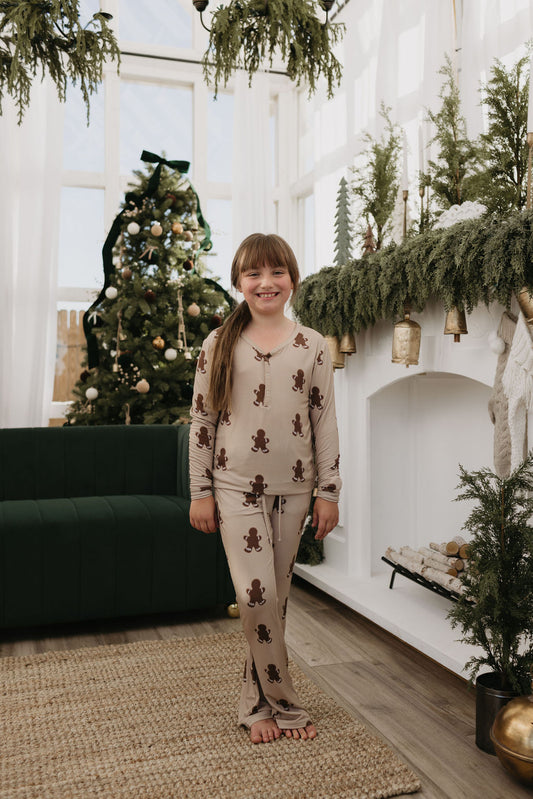 A smiling child stands barefoot in a cozy, holiday-themed room, wearing Pre-Teen Flare Bamboo Pajamas with gingerbread designs from forever french baby. Behind them is a decorated Christmas tree and a mantel adorned with greenery and gold bells.