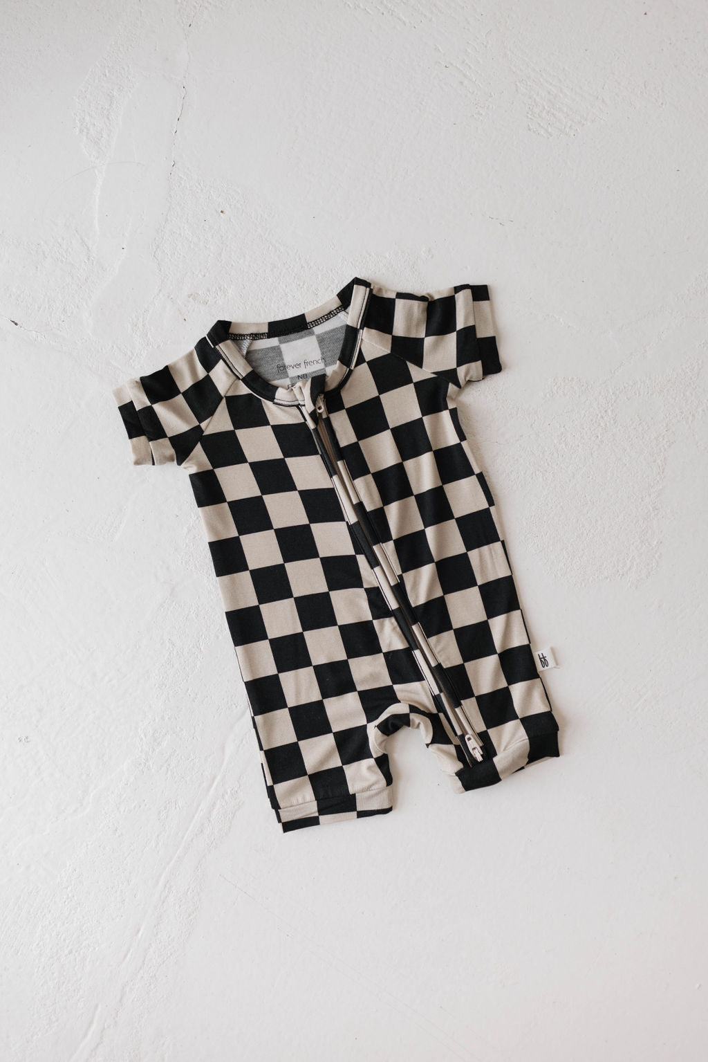 The Shortie Bamboo Zip | Black Checkerboard by forever french baby, a short-sleeved baby romper with a sleek black and white checkerboard pattern, lies flat on a white surface. Made from hypo-allergenic materials, this romper features a front zipper running from the neckline to the crotch area and includes a small tag on the left side seam for added comfort.