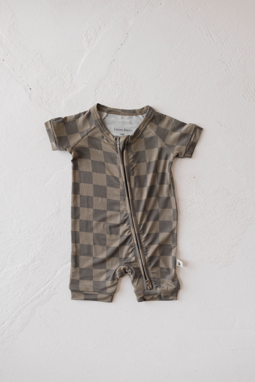 Introducing the Shortie Bamboo Zip in Faded Brown Checkerboard from forever french baby. This baby romper features short sleeves and a convenient front zip closure, designed with alternating light and dark squares in a checkered pattern. Made from breathable bamboo fabric, it ensures softness and comfort while being hypo-allergenic. The product is displayed on a neutral, textured surface for an elegant presentation.