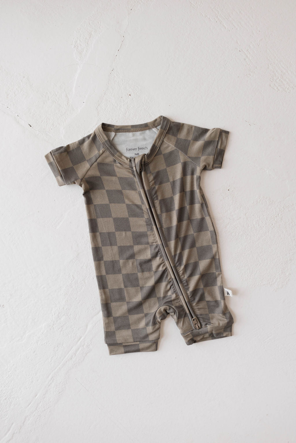 A short-sleeved infant romper, the "Shortie Bamboo Zip | Faded Brown Checkerboard" from forever french baby, is spread out on a light background. Made from breathable bamboo fabric, this hypoallergenic romper features a faded brown checkered pattern and a convenient front zipper.