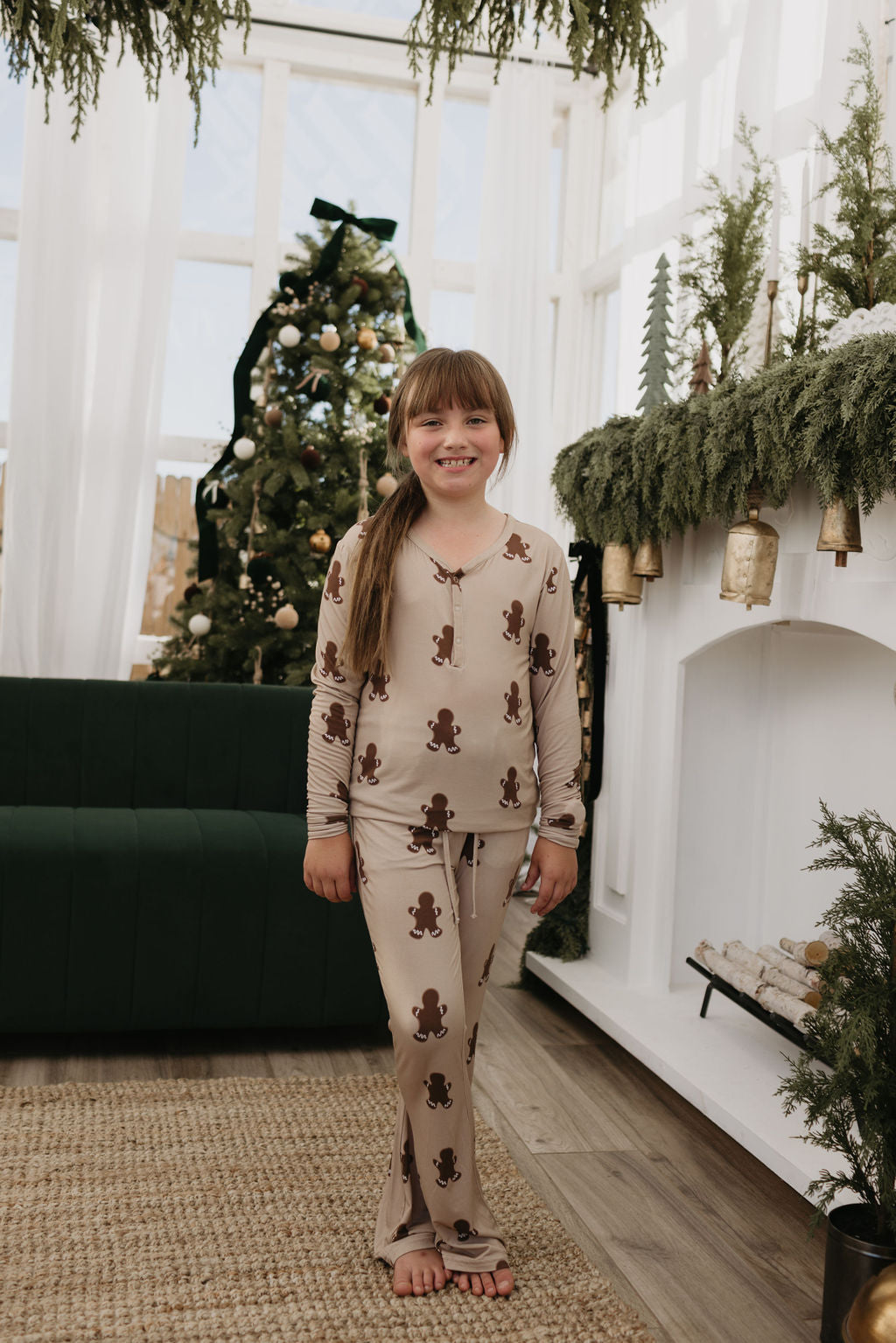 A young girl stands barefoot in a cozy, Christmas-themed room, wearing the charming Pre-Teen Flare Bamboo Pajamas in a gingerbread pattern by forever french baby. Behind her, the Christmas tree glistens beside a fireplace decorated with greenery and ornaments.