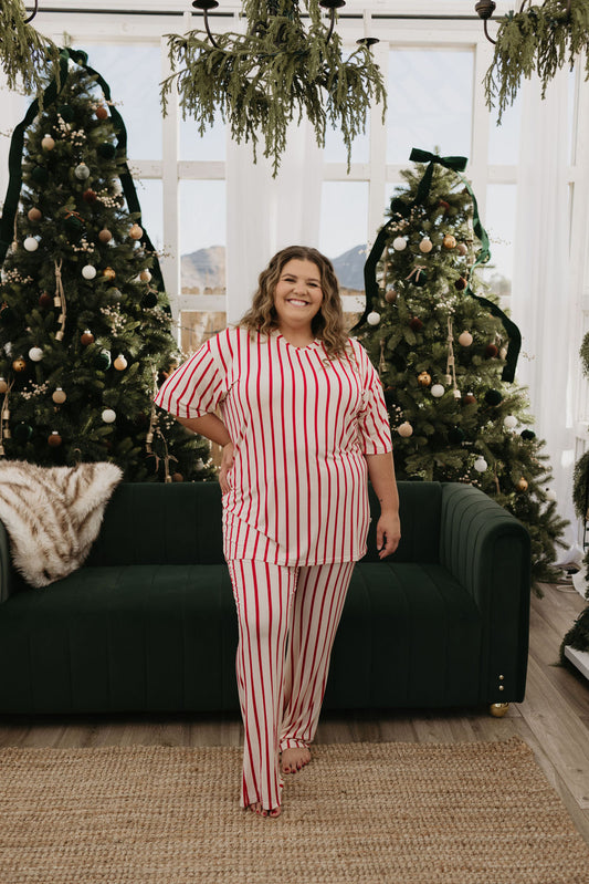 Dressed in a Short Sleeve Women's Bamboo Pajama set, known as the Claus by forever french baby, an individual stands amidst a festive setting featuring three beautifully decorated Christmas trees and a dark green couch. The room is filled with natural light from large windows, creating a cozy holiday atmosphere that perfectly complements the comfort of this oversized look.