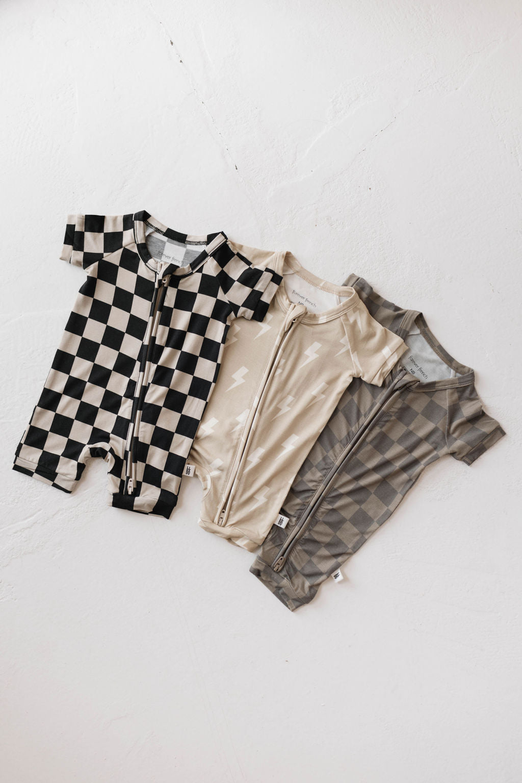 Three baby onesies are neatly laid side by side. The first one is the "Shortie Bamboo Zip | Faded Brown Checkerboard" by forever french baby, featuring a muted checkerboard pattern. The second is tan with a subtle lightning bolt design, and the third is gray with a geometric pattern. All are made from breathable bamboo fabric, equipped with front zippers and short sleeves.