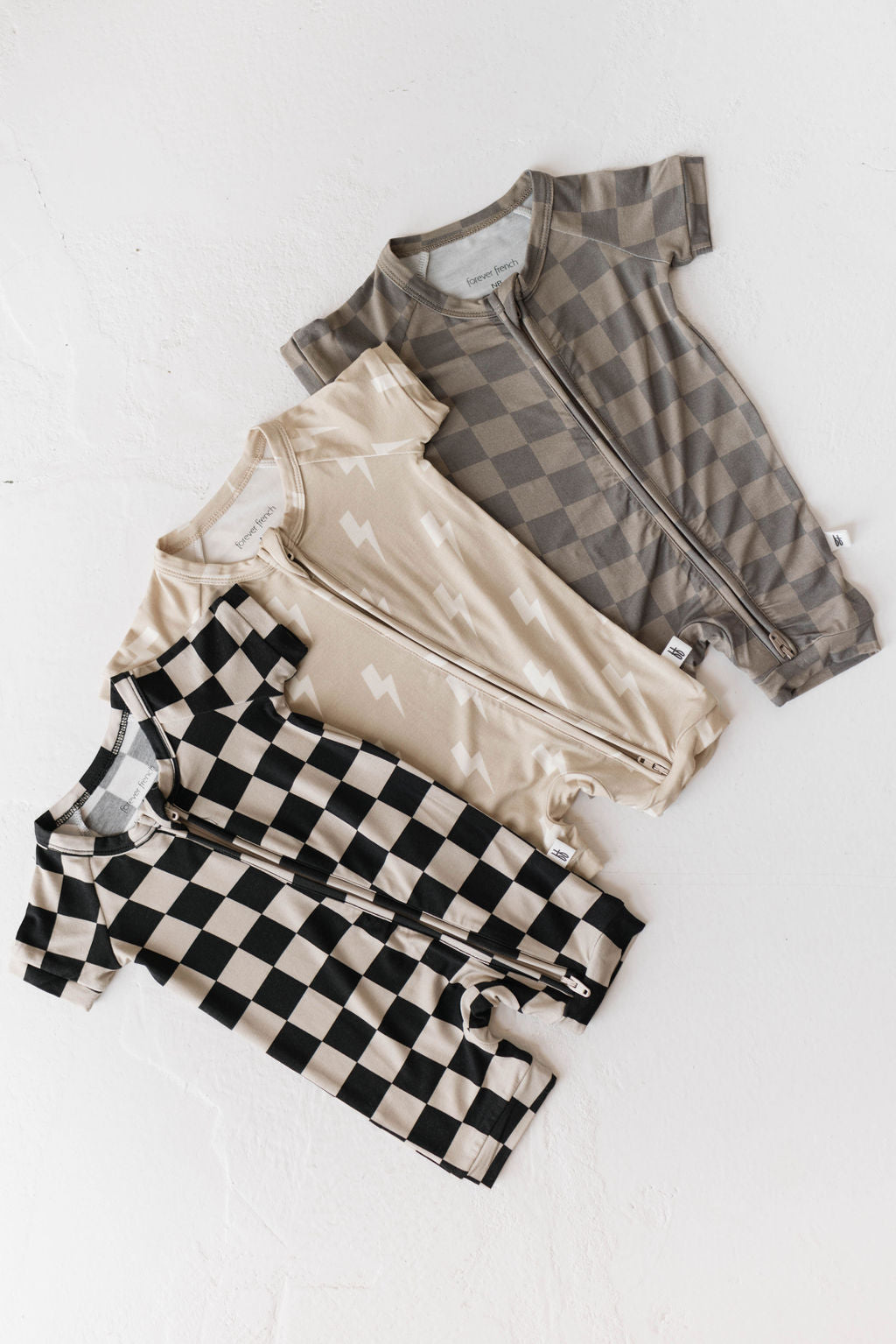 Three baby rompers from Forever French Baby are laid out flat on a light surface. From top to bottom, the baby clothing features a brown checkered pattern, beige with white lightning bolts, and the Shortie Bamboo Zip in Black Checkerboard. Made of soft bamboo pajamas material, all have zippers running down the front.