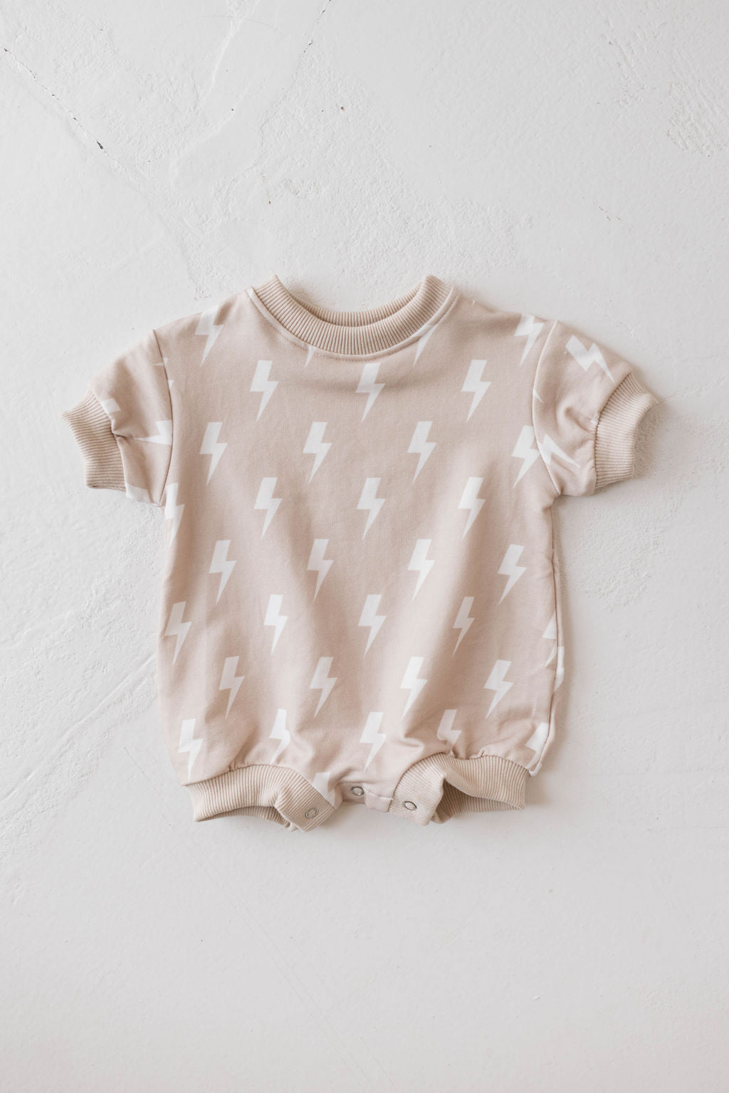 The Sweatshirt Romper from Forever French Baby in Cream Lightning features a pattern of white lightning bolts on muted beige. Made from a soft fabric, it has short sleeves, a round neck, and snap closures at the bottom for easy diaper changes. It is laid out flat on a pale textured surface.