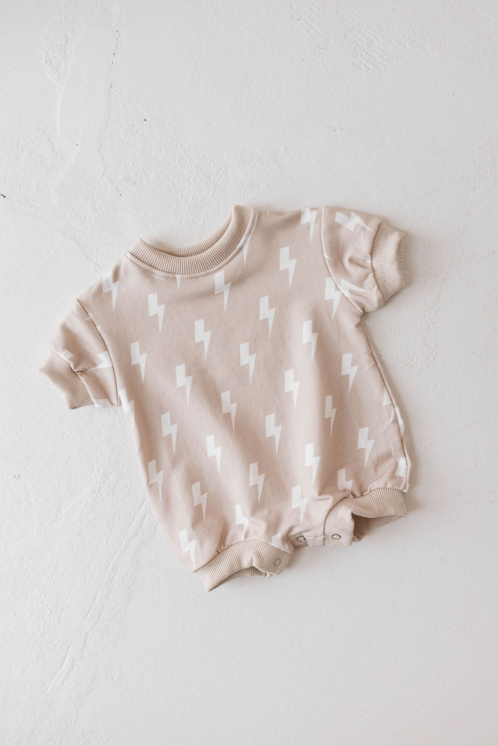 A cream-colored "Sweatshirt Romper | Cream Lightning" with short sleeves and a lightning bolt pattern is laid flat on a white textured background. The onesie, from forever french baby, features ribbed cuffs on the sleeves, neckline, and leg openings, along with snap buttons at the crotch for easy diaper changes in muted tones.