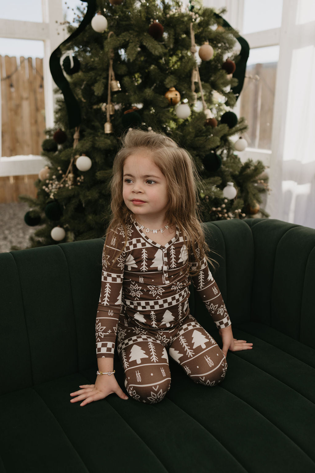 A young girl with long hair sits on a dark green sofa, dressed in Forever Fair Isle Bamboo Two Piece Pajamas by forever french baby, which feature a cozy pattern of white trees and snowflakes. She enjoys the comfort of hypo-allergenic clothing indoors in front of a decorated Christmas tree.