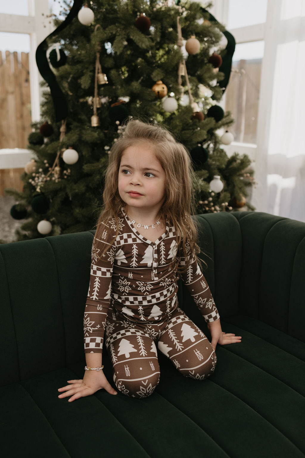 A young child with long hair sits on a dark green couch, wearing cozy Bamboo Two Piece Pajamas in the Forever Fair Isle design by forever french baby. The breathable fabric keeps them comfy as they admire the decorated Christmas tree behind them in a room filled with light from large windows.