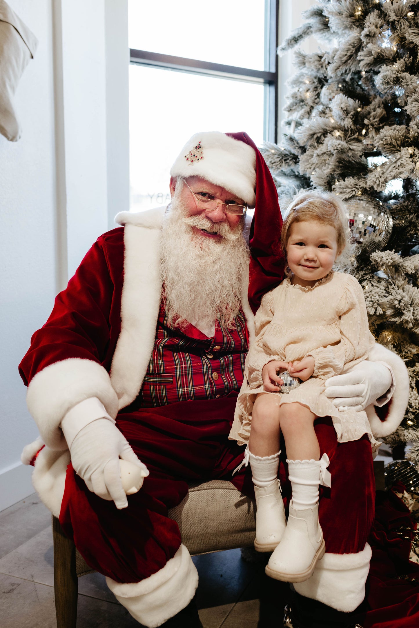 A heartwarming moment is captured by Amanda Riley Photos, showcasing a person dressed as Santa Claus with a smiling young child on their lap in front of a beautifully decorated Christmas tree. The child, donning a pale dress and white boots from the Forever French Baby collection, looks joyfully at the camera. This festive scene perfectly encapsulates the spirit of Amanda Riley Photos' special collaboration: Santa Photos | Forever French Baby x Amanda Riley Photos.