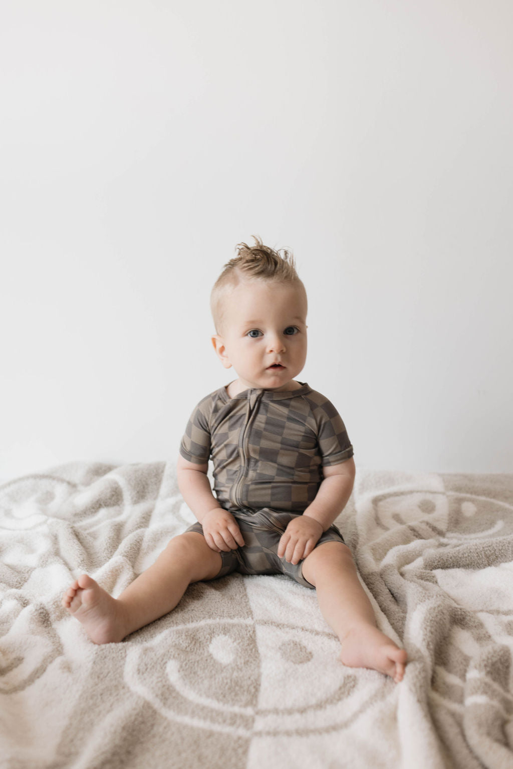 A toddler with blonde hair sits on a white and beige blanket with a subtle pattern. The child is wearing the Shortie Bamboo Zip in Faded Brown Checkerboard by forever french baby, an outfit that is both breathable and hypo-allergenic, appearing to be looking slightly off-camera. The background is plain and white.