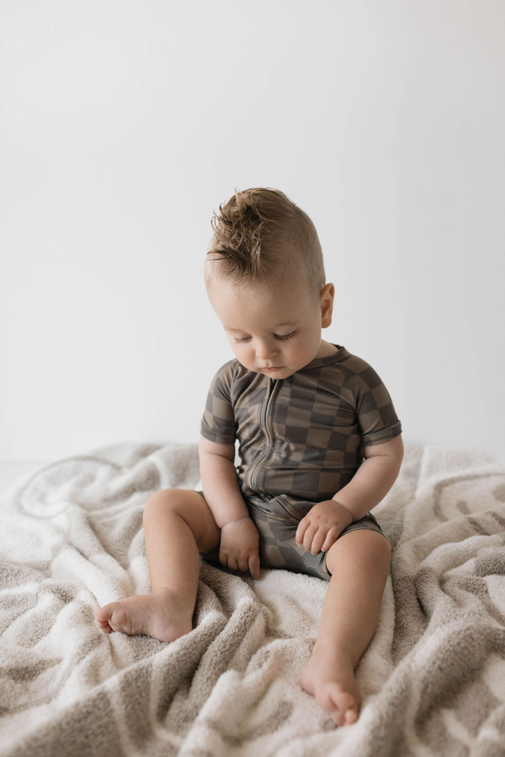 A baby with light hair styled in a fauxhawk, wearing the "Shortie Bamboo Zip | Faded Brown Checkerboard" onesie by forever french baby, sits on a patterned blanket. They look downward with their hands resting on their legs against a neutral and minimalistic background.